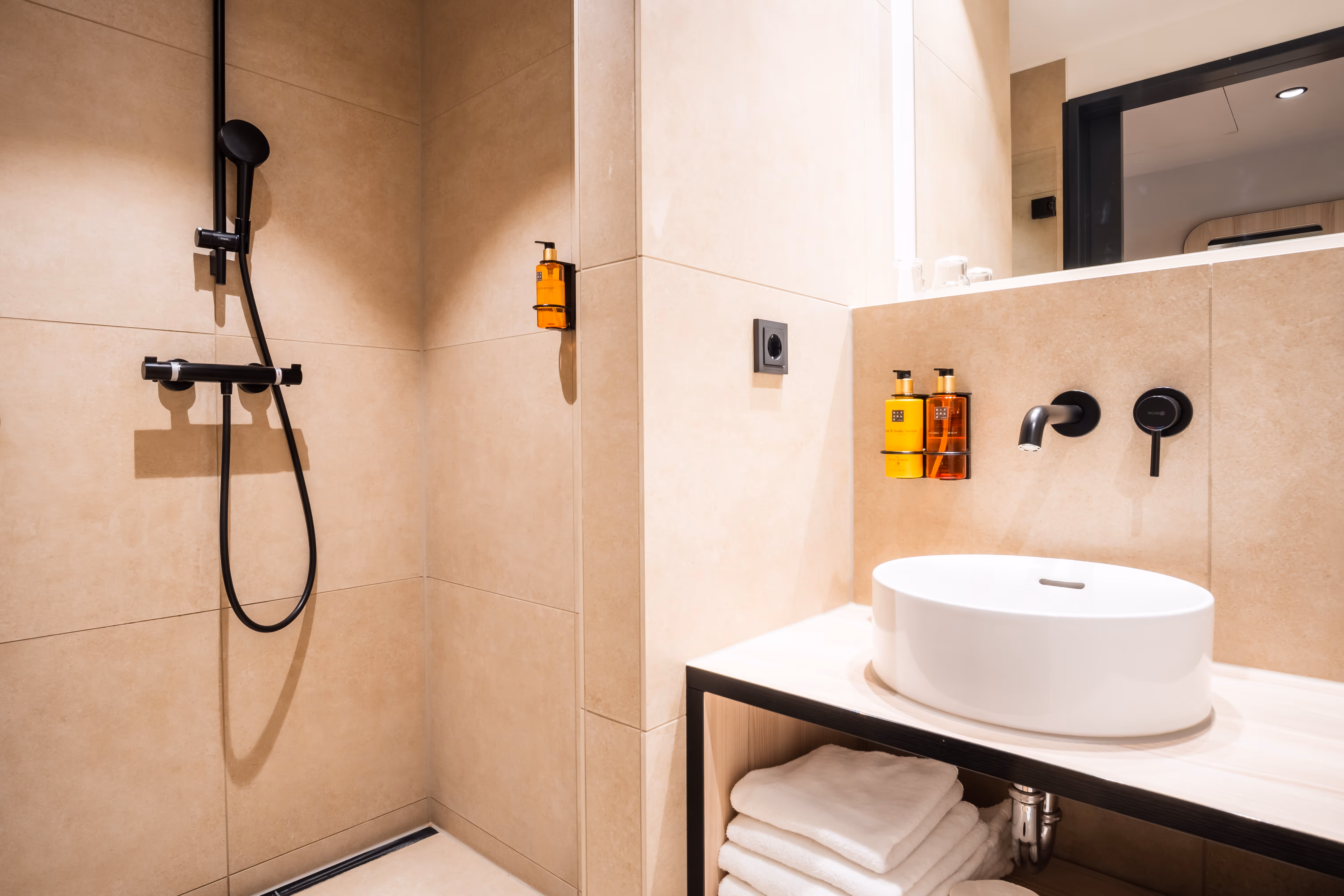 Modernes Badezimmer mit beige Fliesen, schwarzer Handbrause, rundem weißen Waschbecken und Stapel weißer Handtücher.