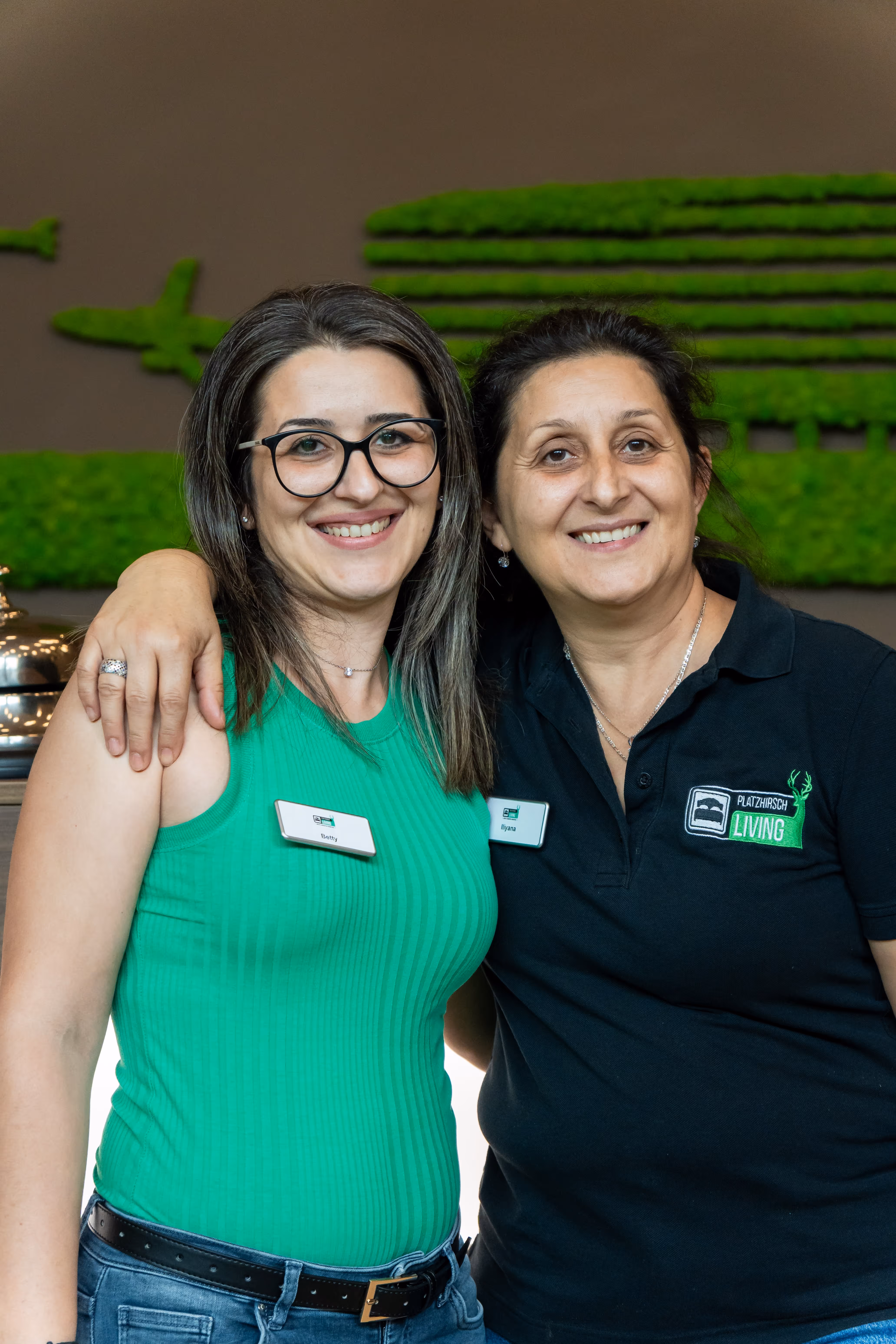 Zwei lächelnde Frauen, die Arme umeinander gelegt haben, eine in grünem Oberteil mit Brille, die andere in schwarzem Poloshirt mit Firmenlogo.