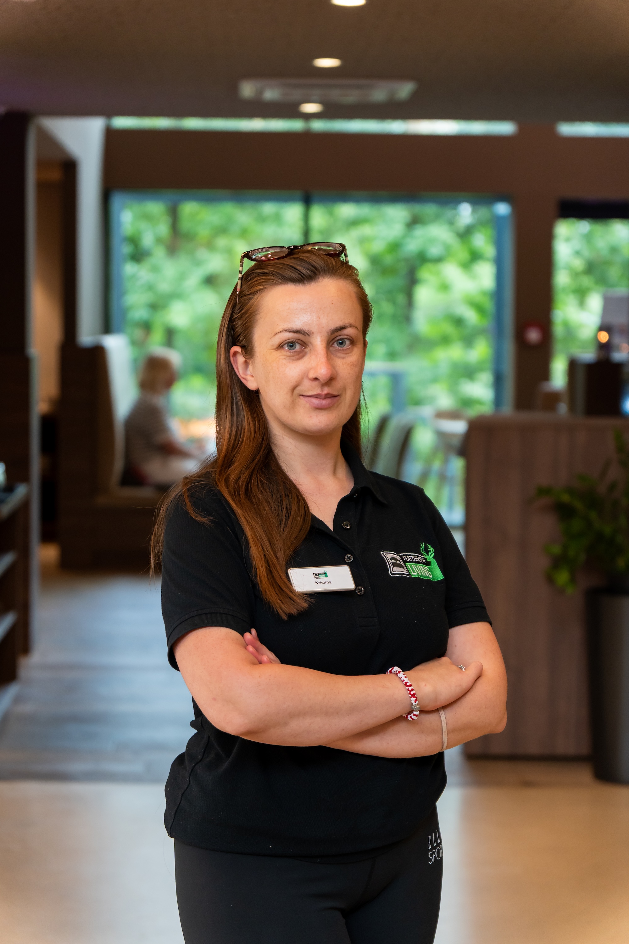 Frau mit dunklem Poloshirt und Namensschild steht mit verschränkten Armen in einem modernen Raum mit großen Fenstern und grüner Aussicht.