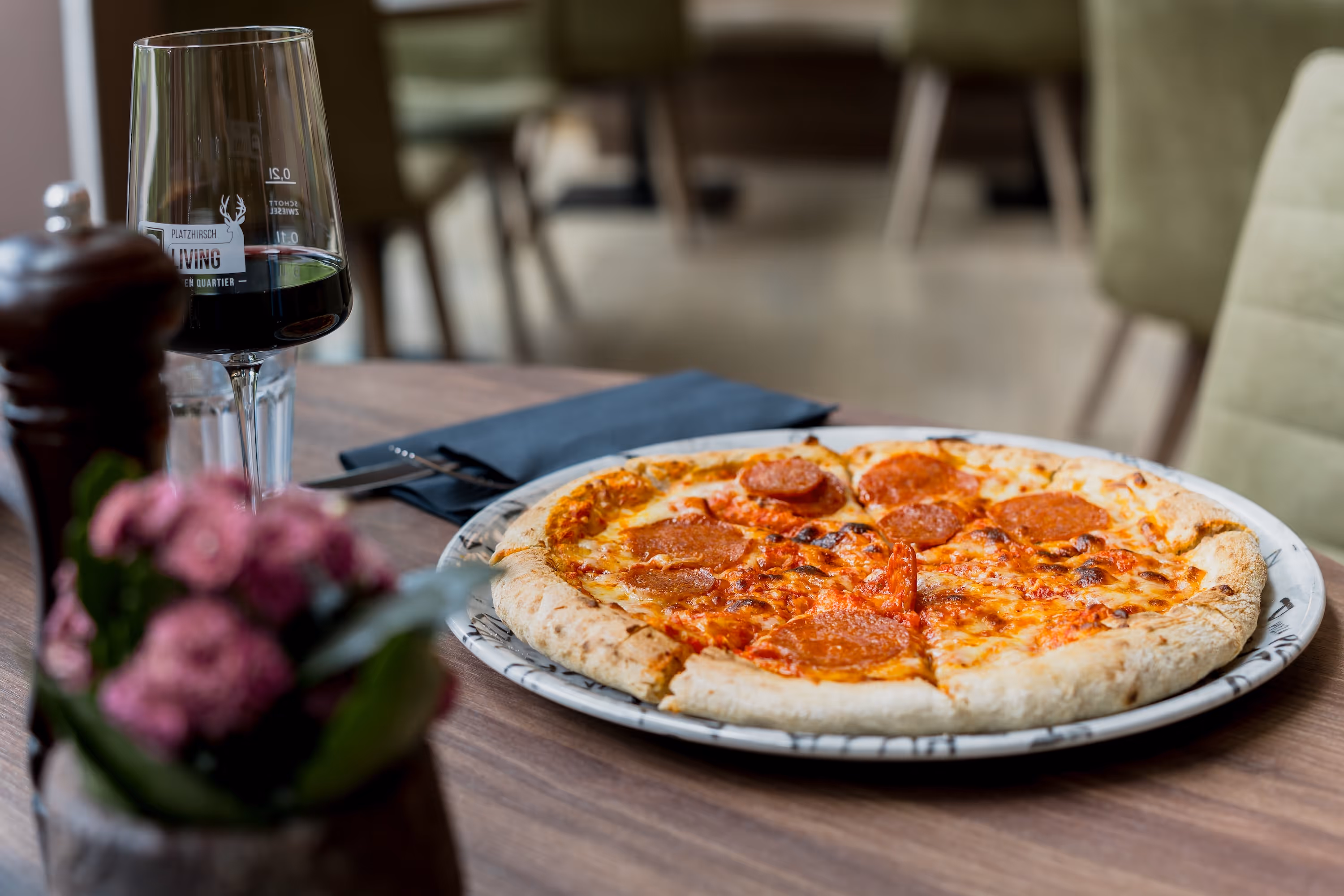 Pepperoni-Pizza auf einem Teller auf einem Holztisch neben einem Glas Rotwein und einer Vase mit rosa Blumen.
