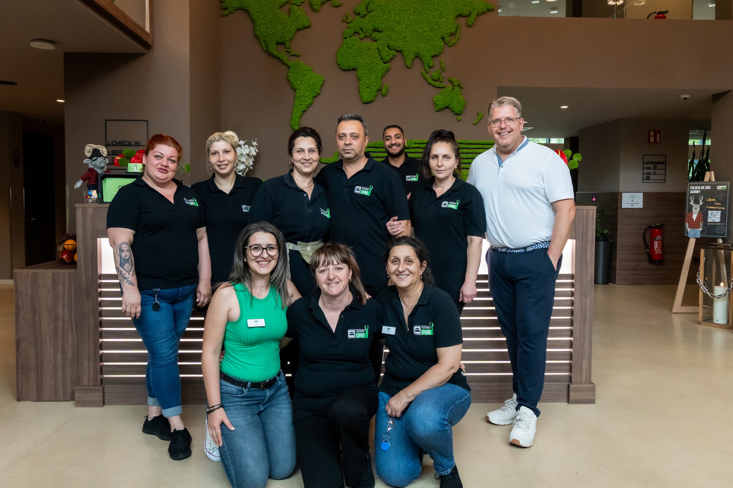 Gruppenfoto von neun Personen, hauptsächlich in schwarzen Firmenpoloshirts, vor einer Rezeption mit einer grünen Weltkarte an der Wand.