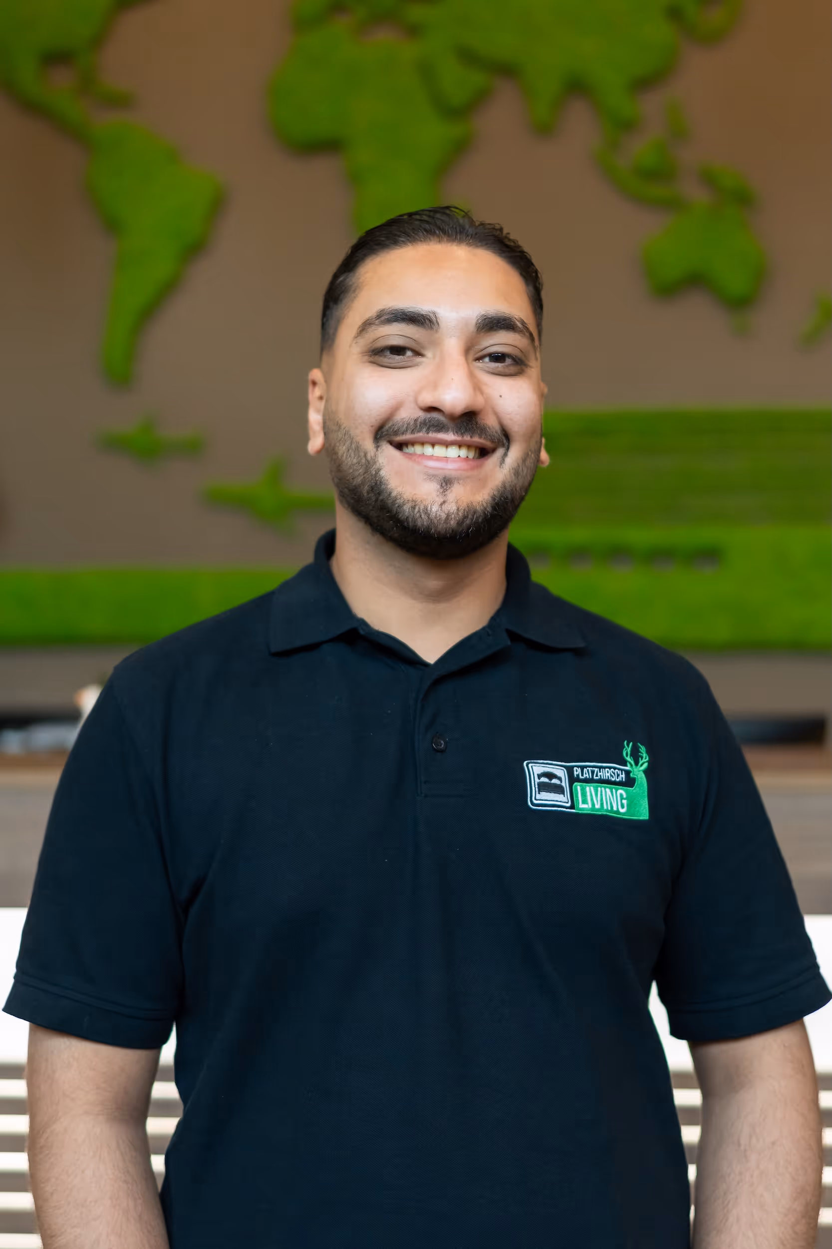 Lächelnder Mann mit dunklen Haaren und Bart trägt ein schwarzes Polohemd mit Platzhirsch Living Logo vor einer grünen Weltkarte aus Moos.