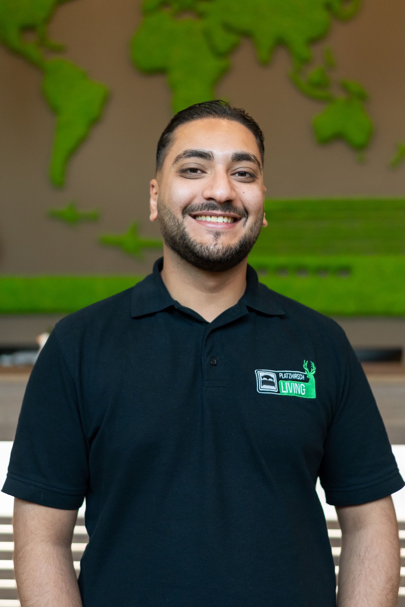 Lächelnder Mann mit dunklen Haaren und Bart trägt ein schwarzes Polohemd mit Platzhirsch Living Logo vor einer grünen Weltkarte aus Moos.