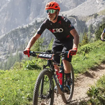 Mountain biker wearing an orange helmet and black cycling gear riding on a trail with rocky cliffs and greenery in the background.