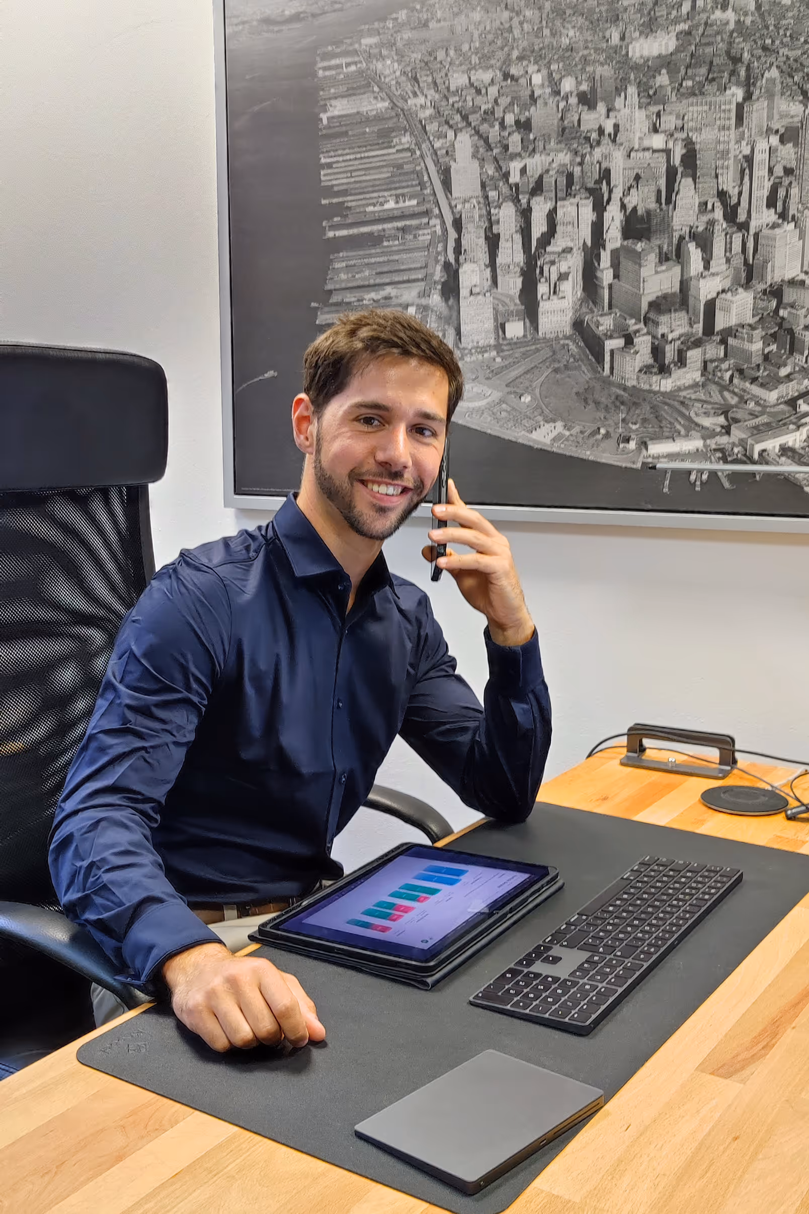 Felix Krotz sitzt am Schreibtisch vor Tablet und Telefon und lächelt