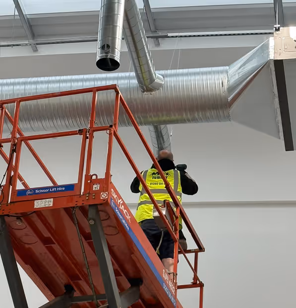 Man wearing a yellow safety vest standing on a red scissor lift working near large silver ductwork inside a building.