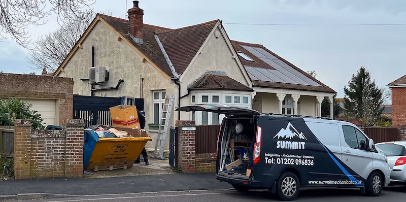 Residential house with solar panels on the roof, a large yellow skip filled with debris, and a black Summit service van parked in front with the back door open.