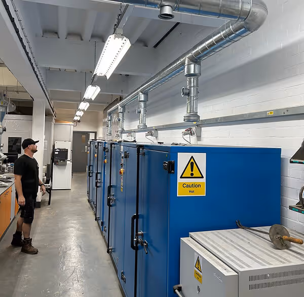 Summit Mechanical engineer wearing black clothing and brown boots standing in an industrial room next to large blue machinery with caution signs and overhead metal ventilation pipes.