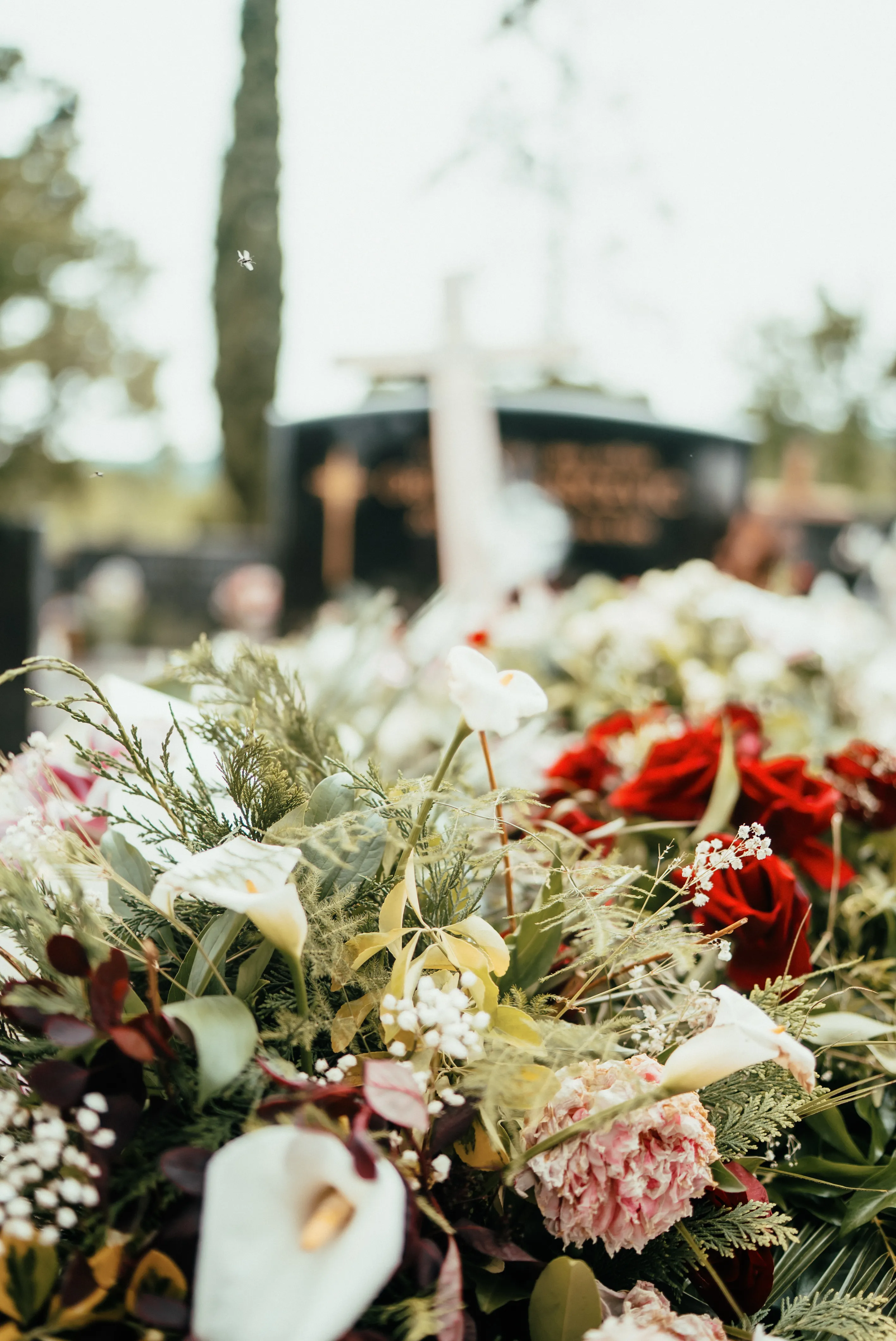 Close-up of mixed floral funeral arrangement with white lilies, red roses, greenery, and blurred tombstones in the background.