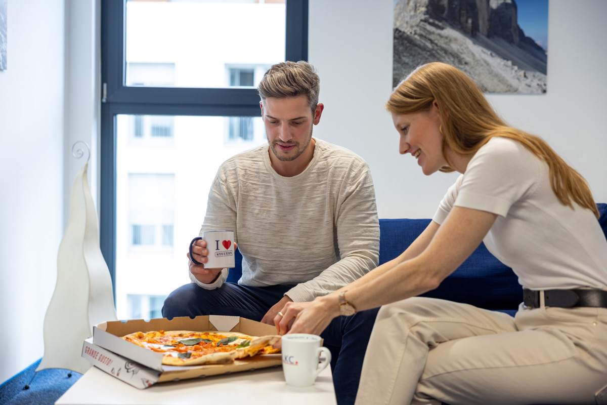 Zwei Personen im Büro beugen sich über eine Pizza auf dem Tisch.