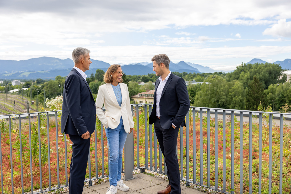 Drei Personen im Business-Outfit im Gespräch draußen auf einer Dachterrasse.