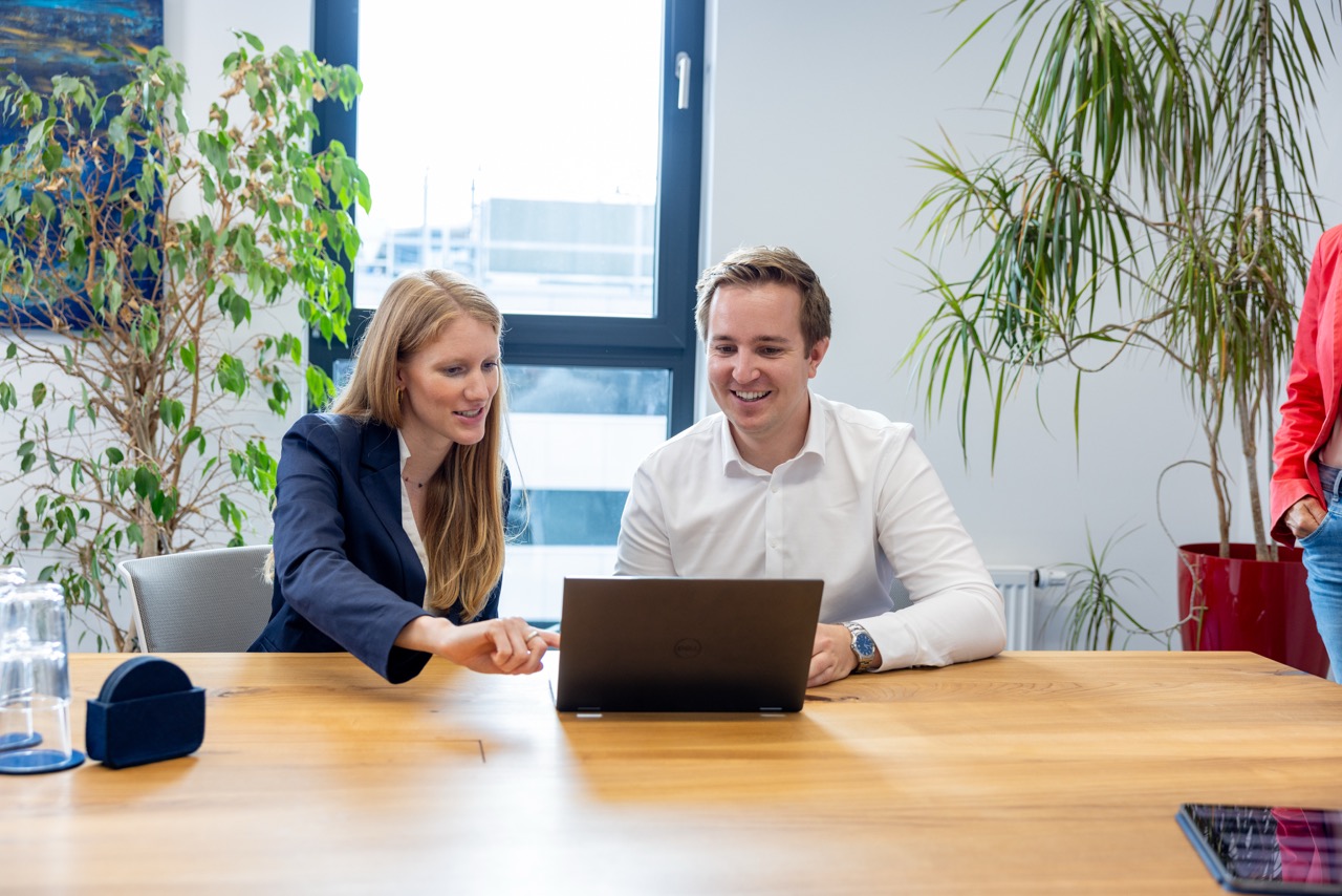 Zwei Personen bei einer Besprechung am Tisch mit Laptop, Fenster im Hintergrund.