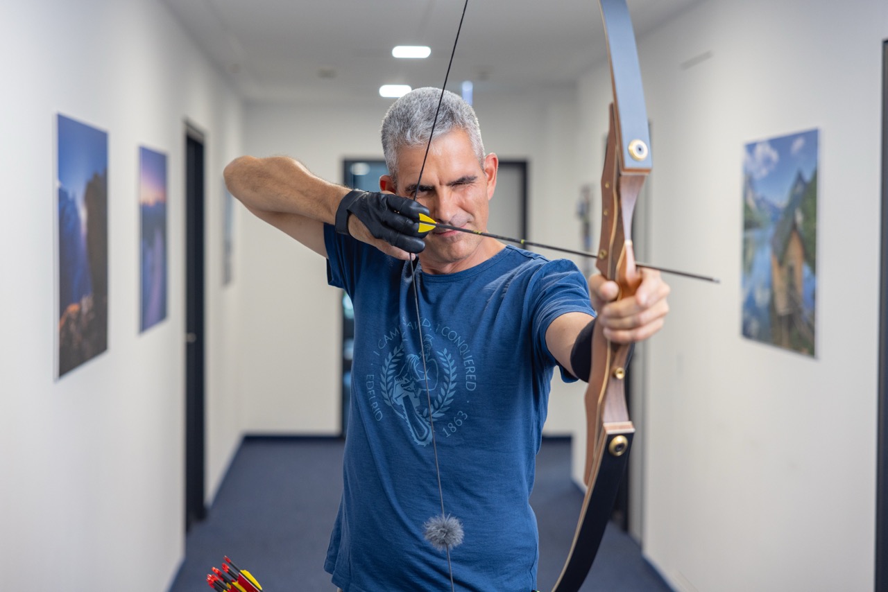 Mann im Flur spannt Pfeil und Bogen und lächelt.