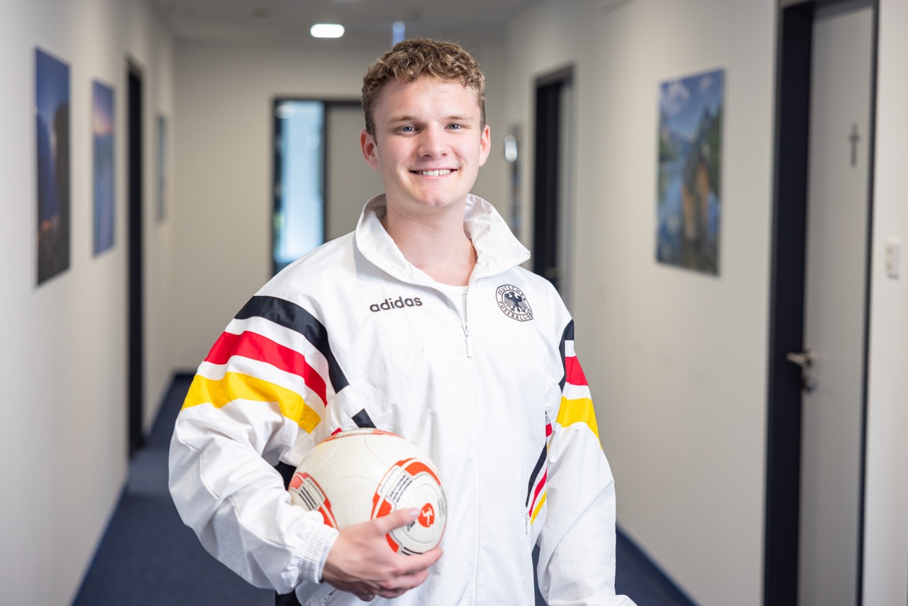 Mann im Flur trägt Fußballjacke, hält einen Fußball und lächelt.