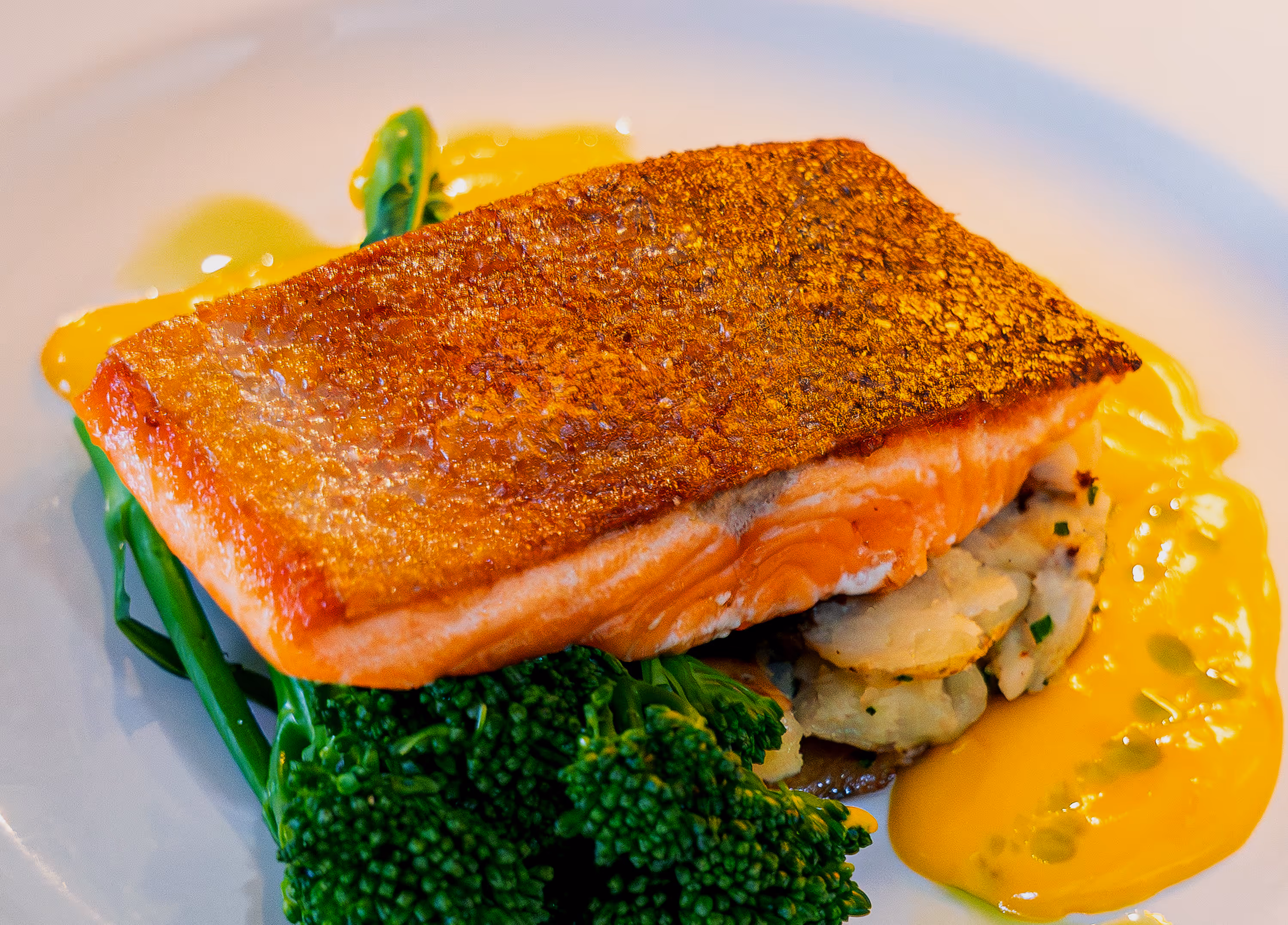 Pan-seared salmon fillet served on sautéed potatoes and broccolini with a side of yellow sauce on a white plate. Served from a pub restaurant 