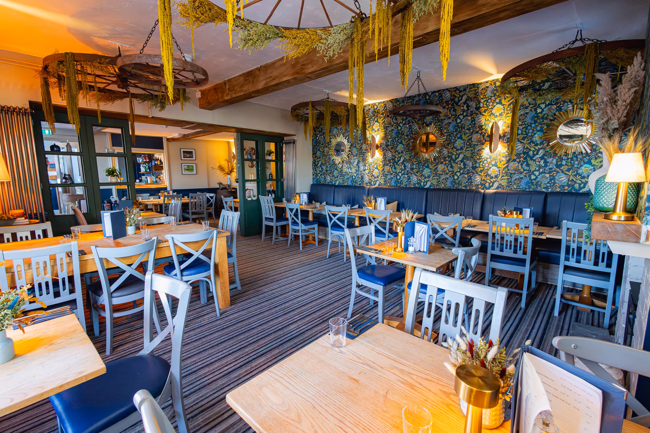 Inside of our Evesham pub restaurant interior with wooden tables, grey chairs, floral wallpaper, hanging dried plants, and soft lighting.