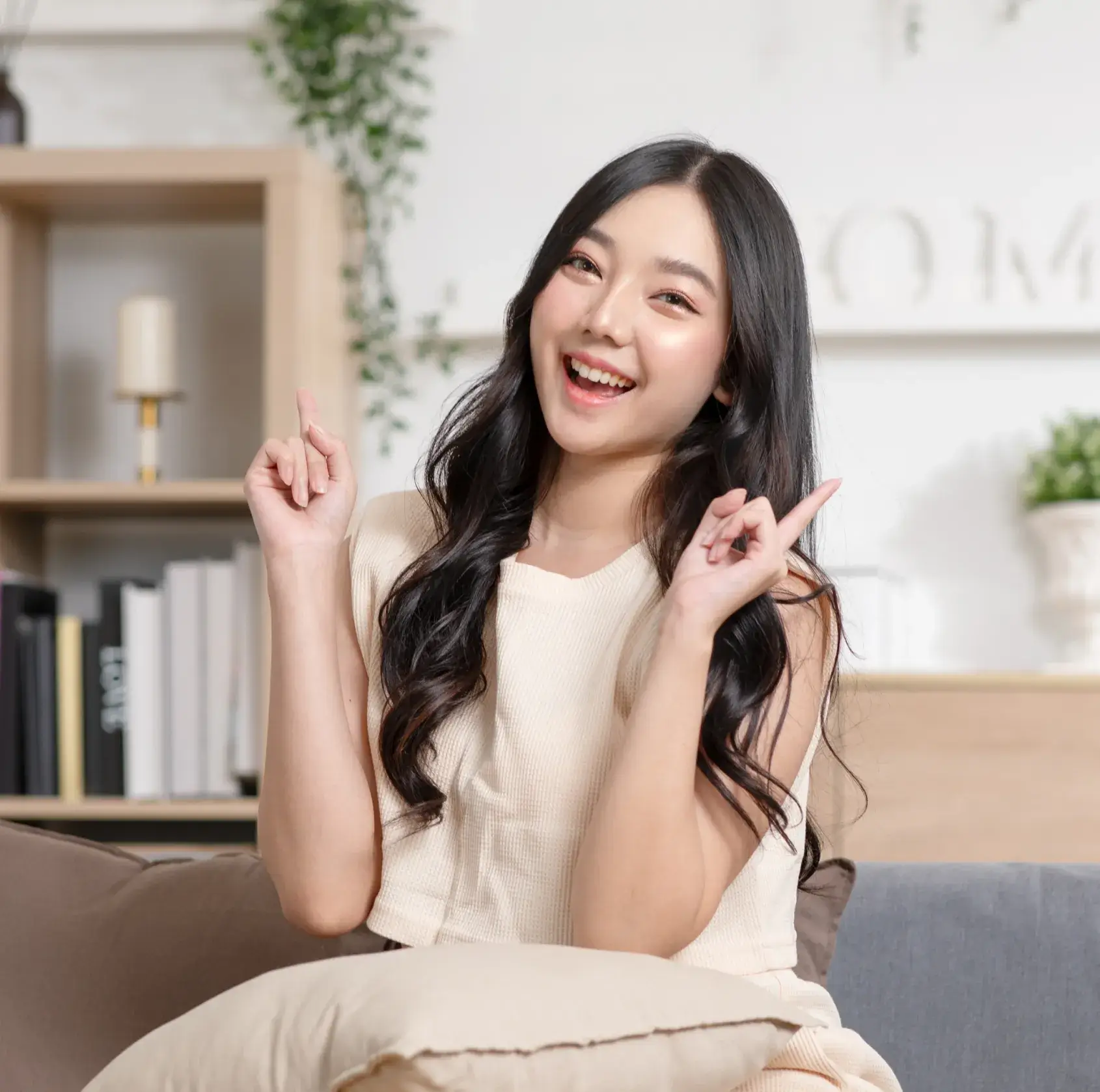 A woman sitting on a couch, smiling and pointing upwards with both index fingers.
