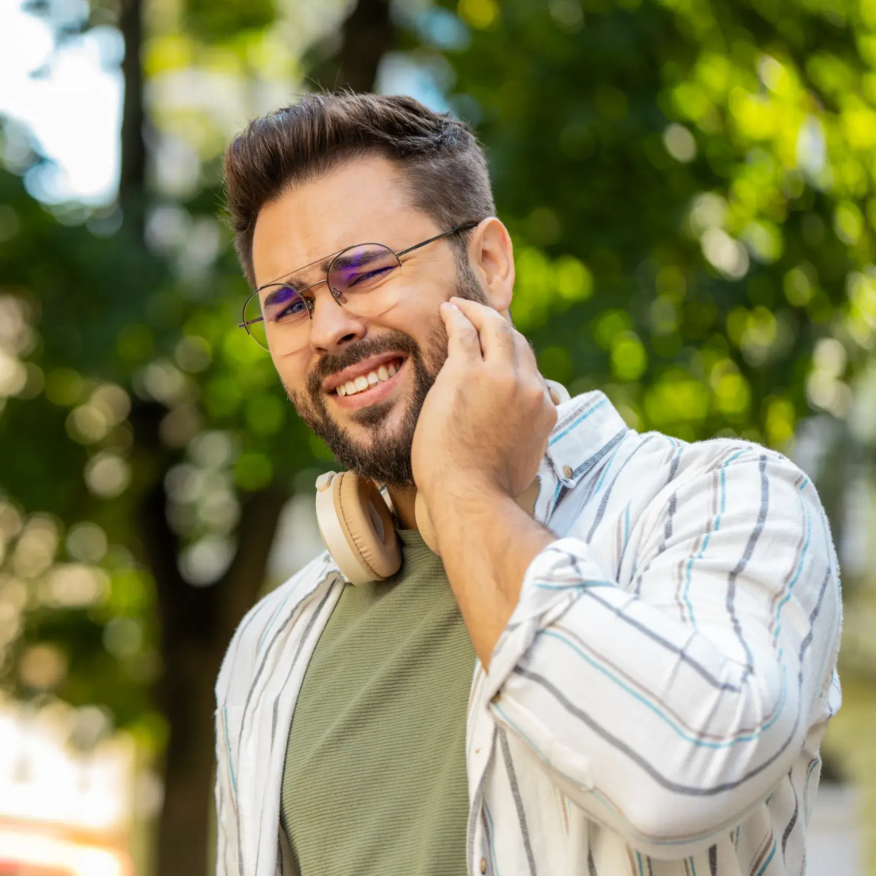Man with headphones smiling, touching his face, standing outdoors in a striped shirt.