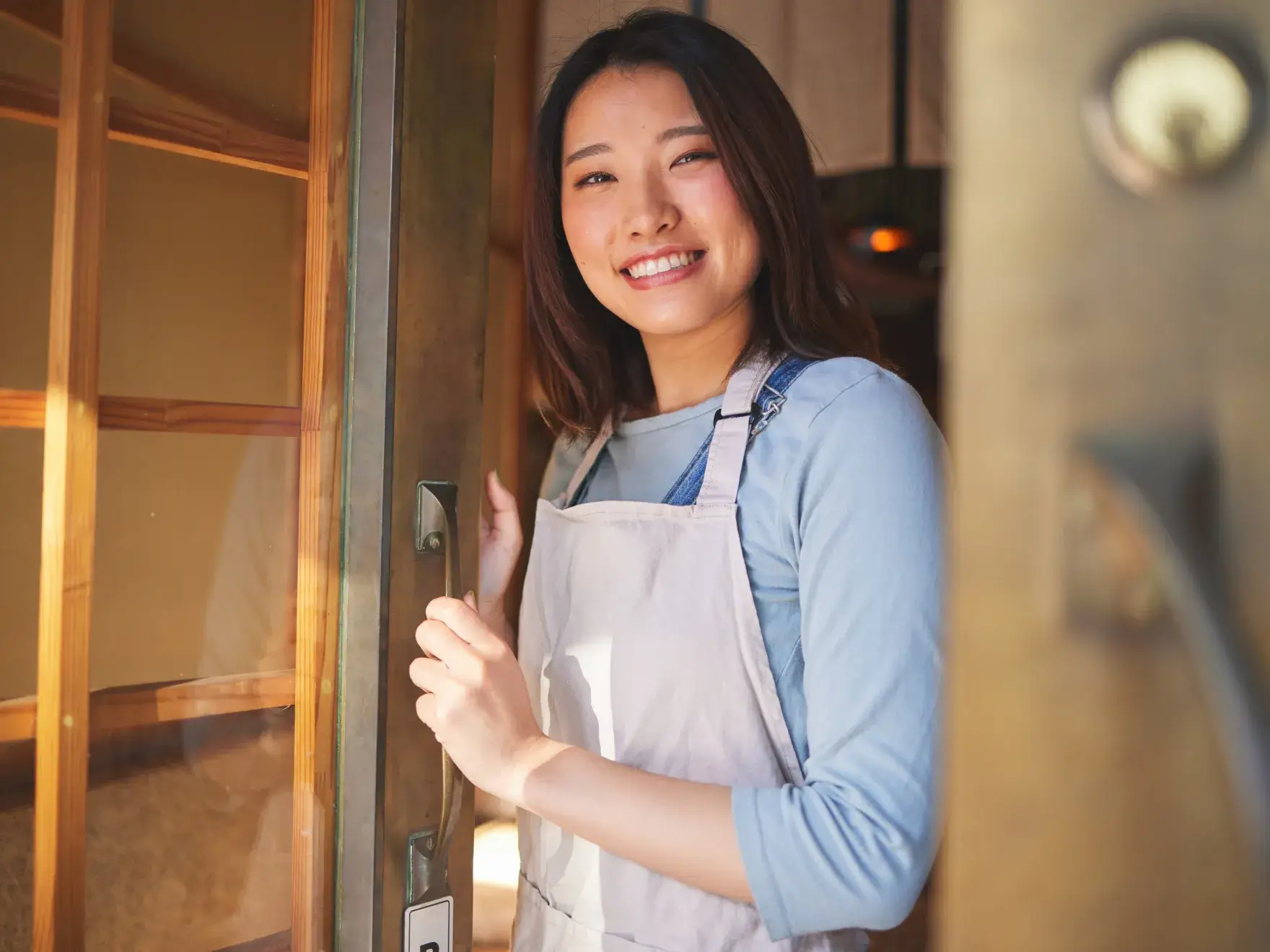 A woman wearing an apron smiles while standing at a doorway, holding the door open.