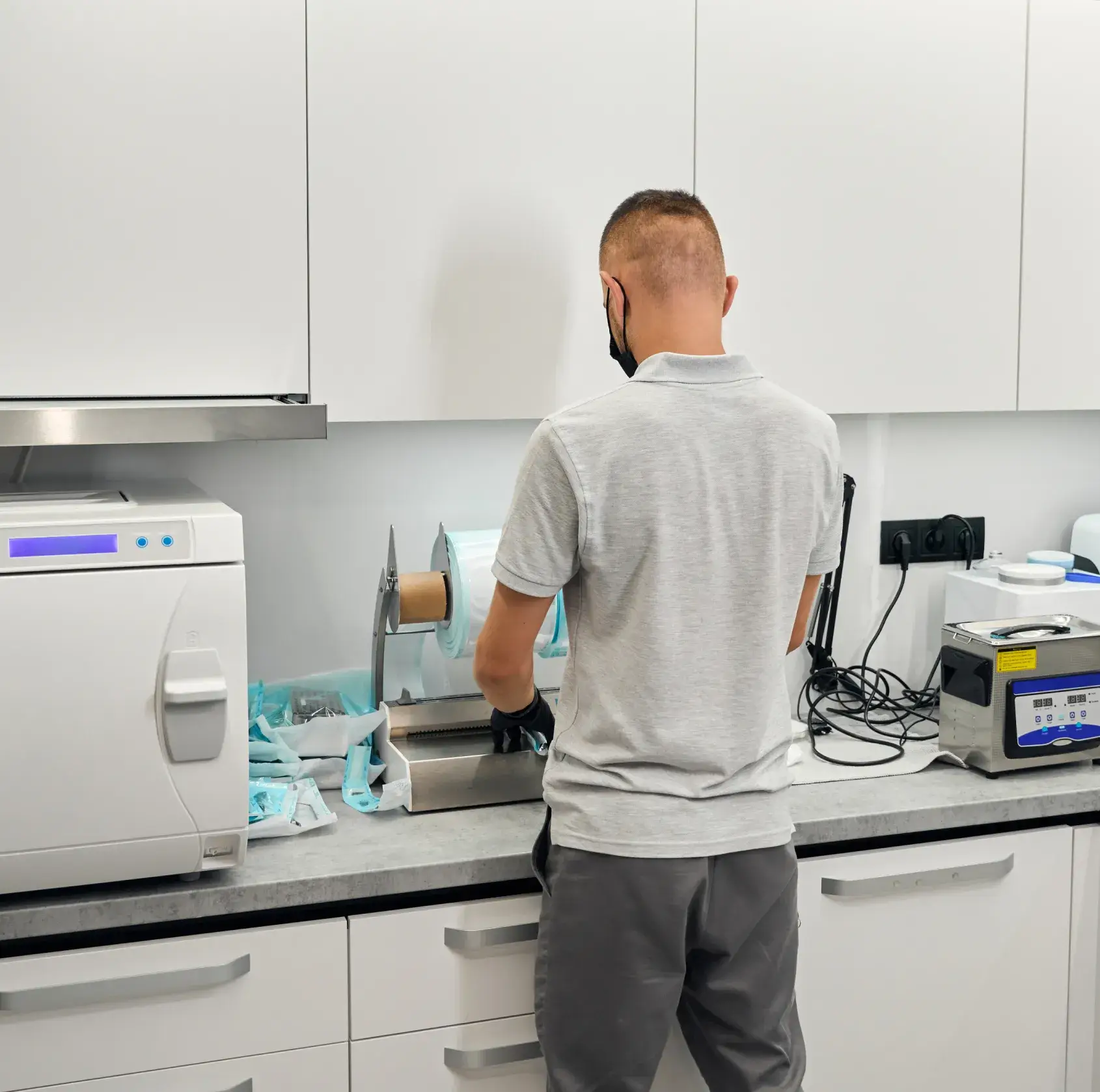 A person wearing a mask operates equipment in a white laboratory or medical setting.