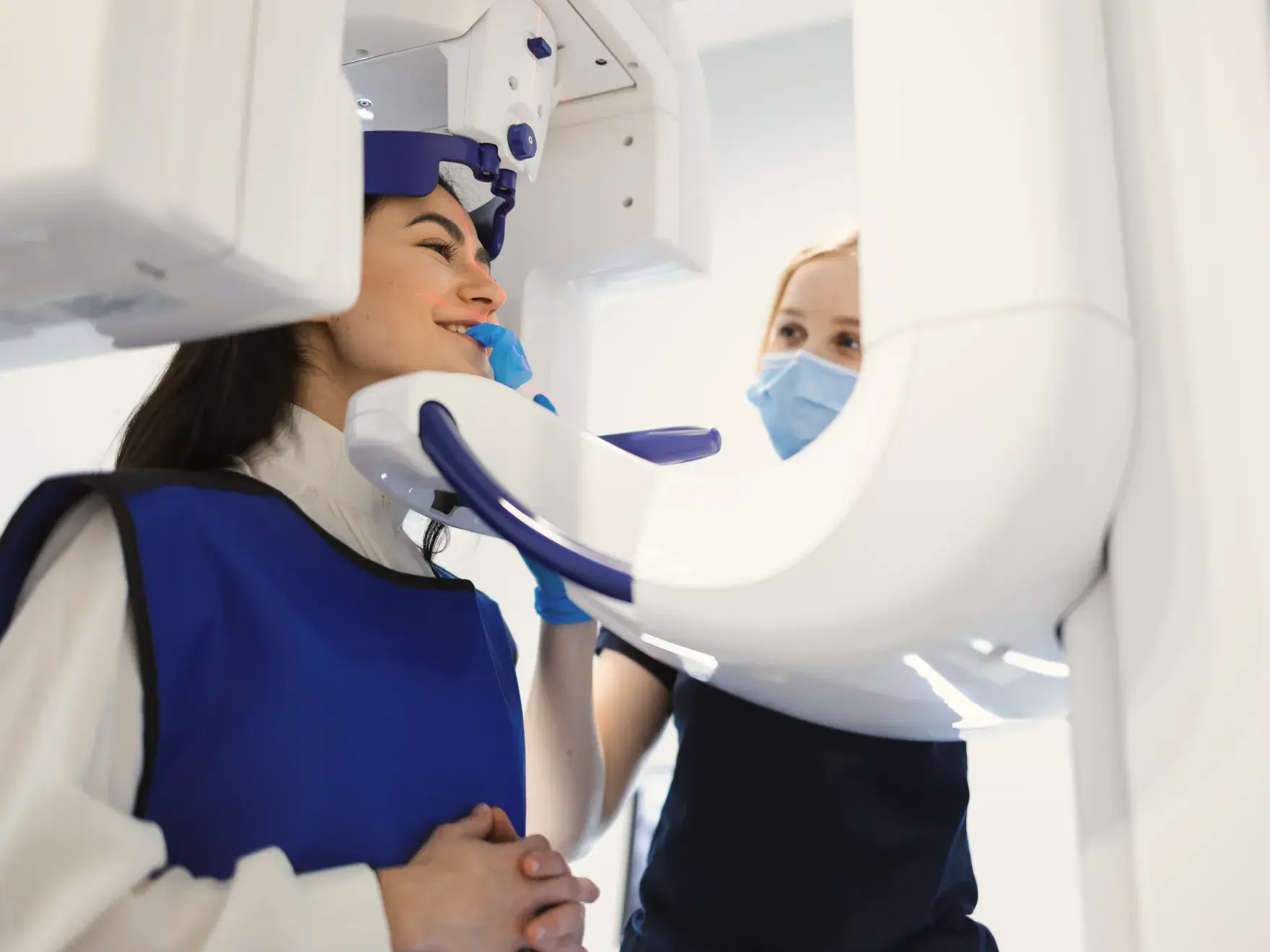 A woman receives a dental X-ray while a dental assistant oversees the procedure, both wearing protective gear.