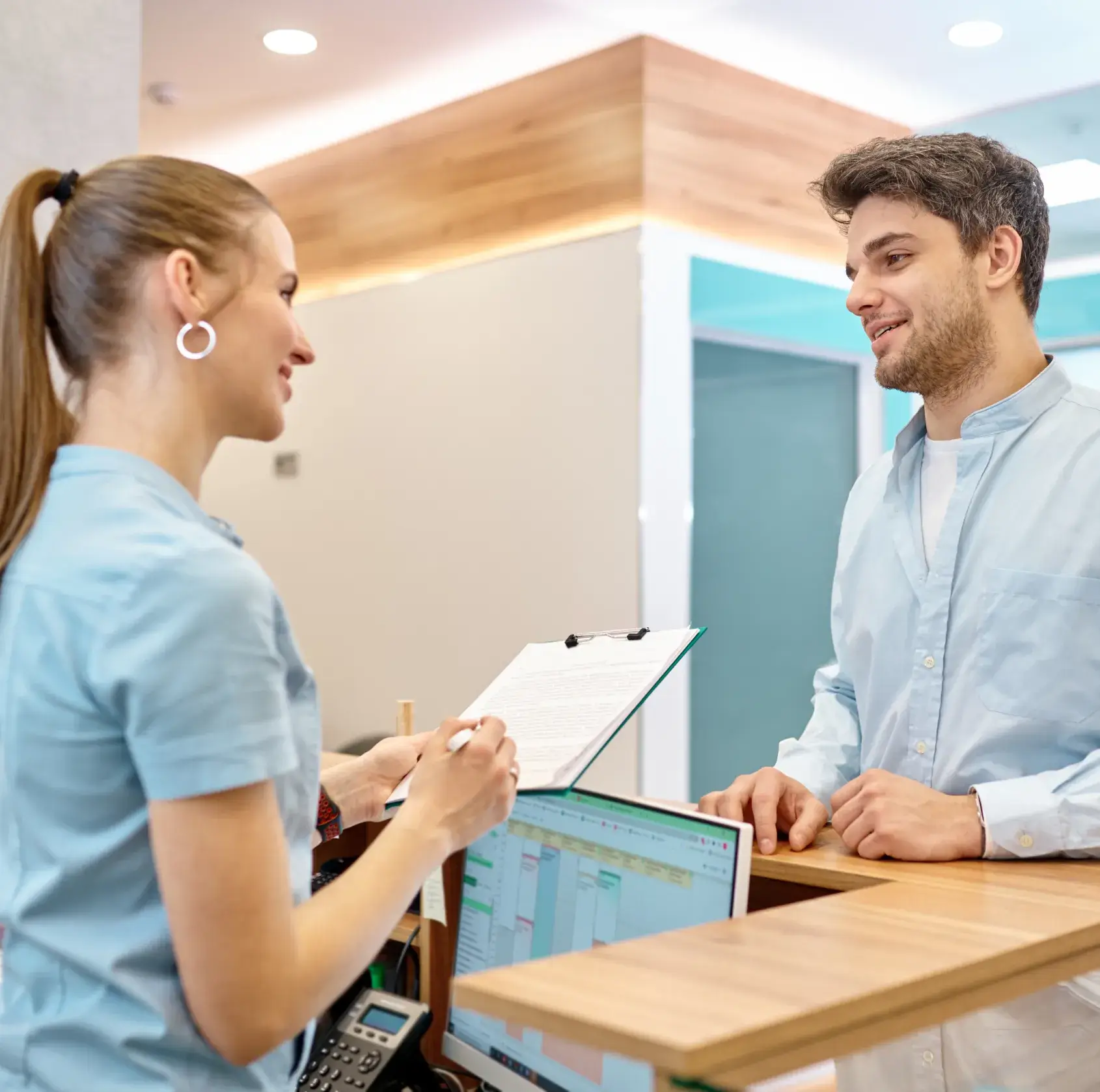 Woman in scrubs holds a clipboard, talking to a man at a reception desk.