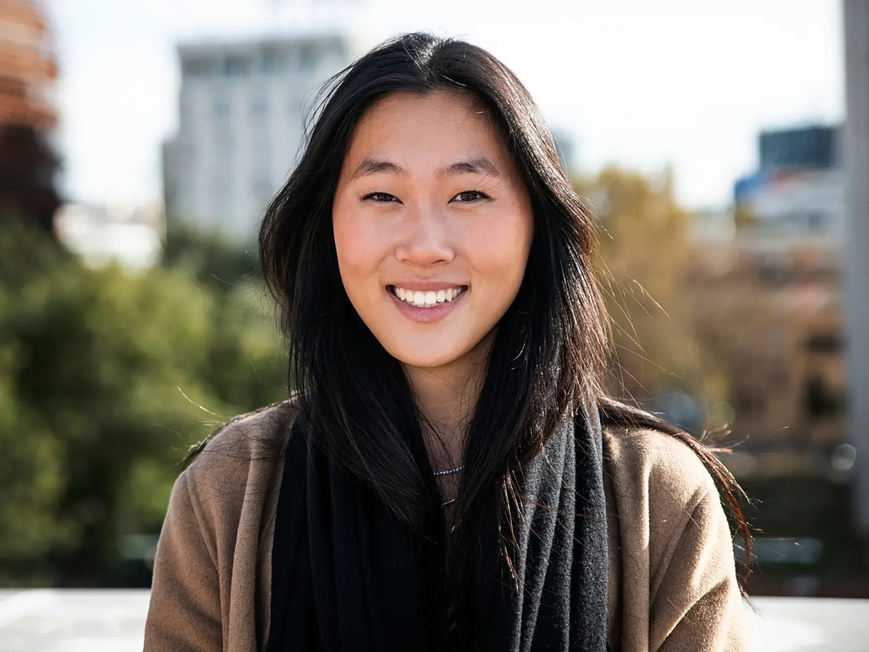 A person smiling outdoors with trees and buildings in the background.
