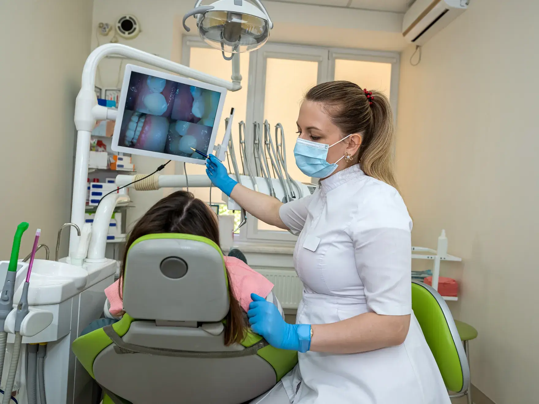 A dentist in protective gear explains a dental procedure to a seated patient using a monitor.