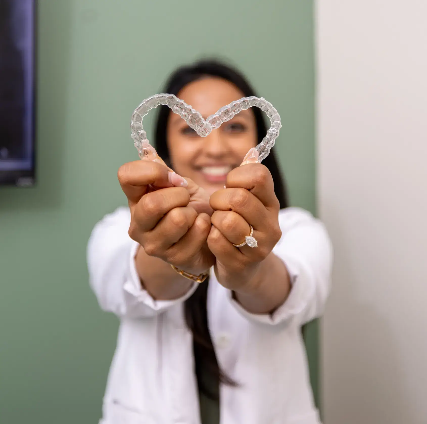 Person holding two clear aligners forming a heart shape with hands stretched forward, smiling in the background.