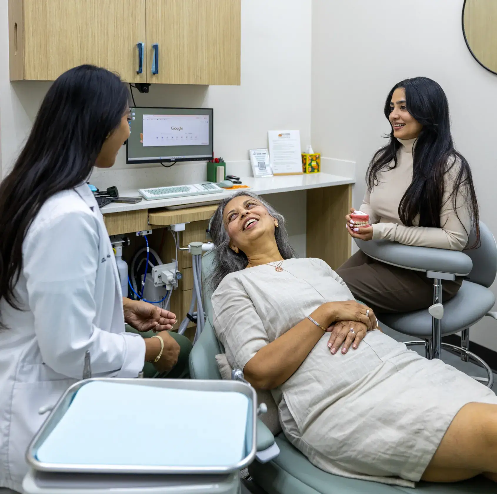 Dental patient reclining in chair smiling while dentist and assistant discuss dental model in office.