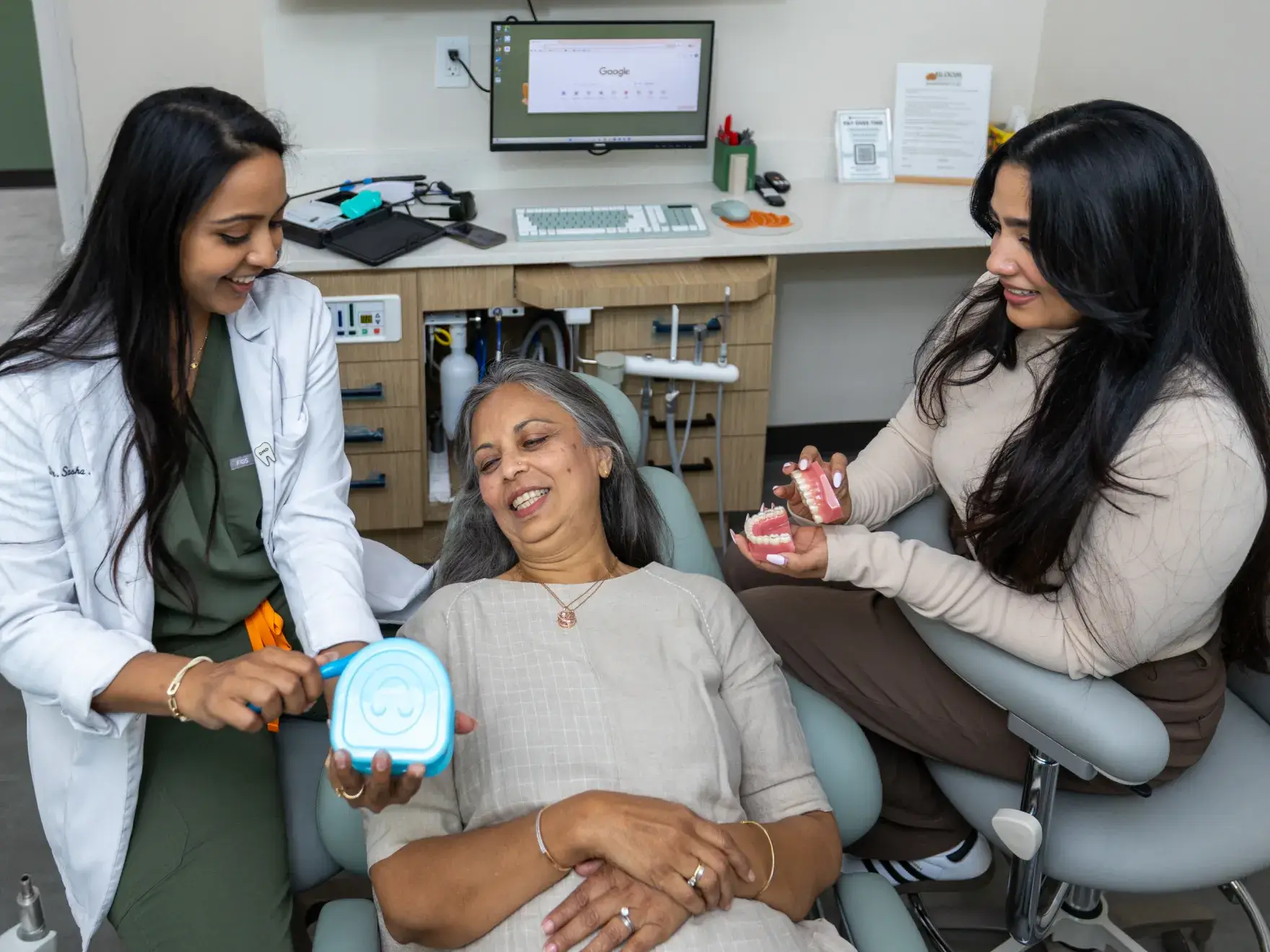 Dentist in white coat showing dental mold and another woman holding a model of teeth while an older woman reclines in a dental chair smiling.