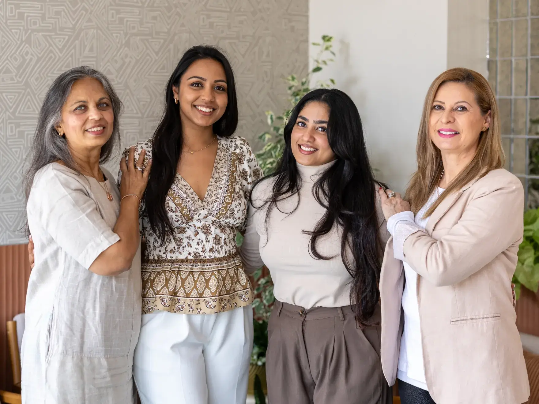 Four women of different ages standing indoors, smiling, with their hands gently resting on each other's shoulders.