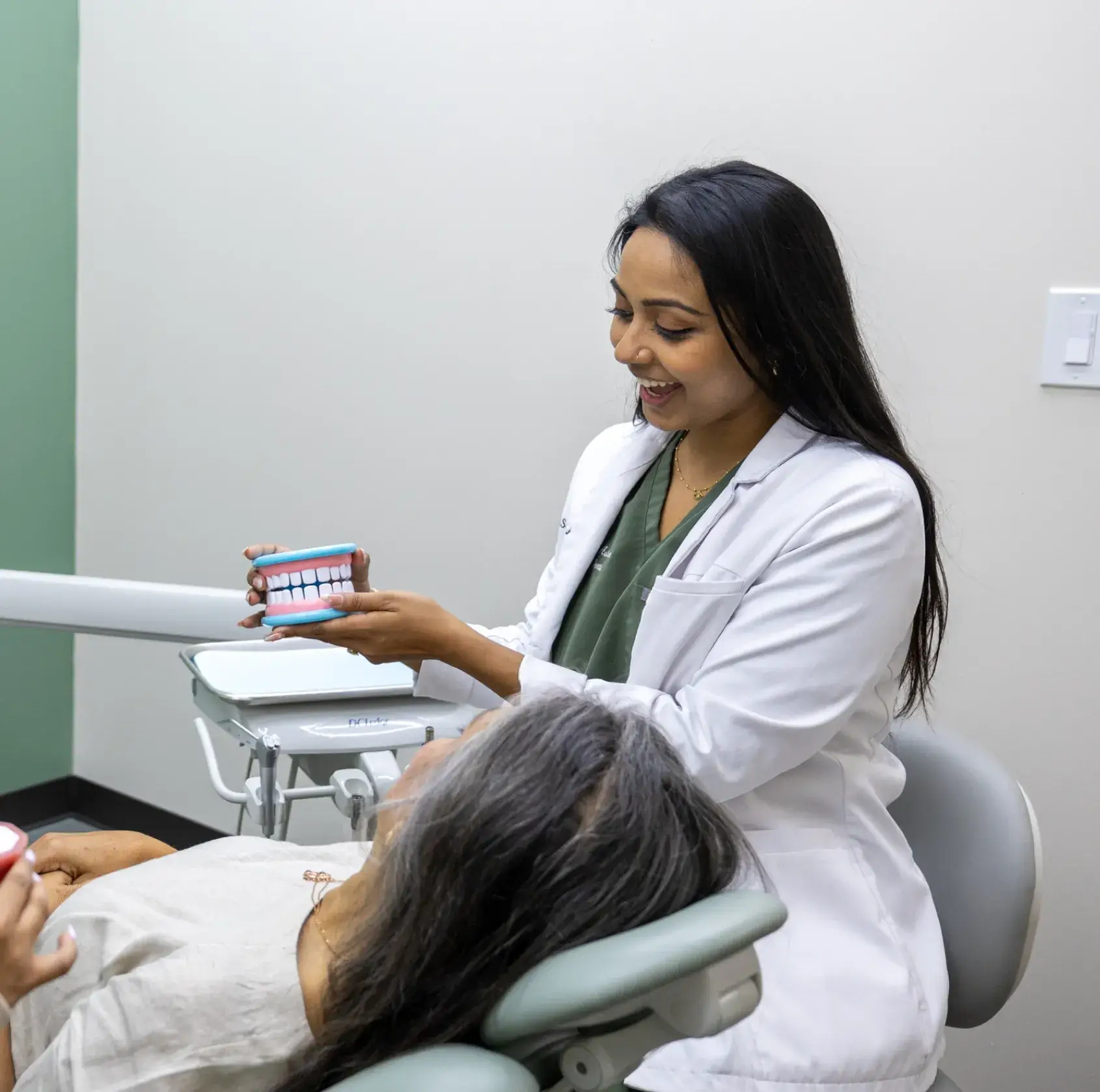 Dentist in a white coat demonstrating dental care using a large teeth model to a patient in a dental chair.