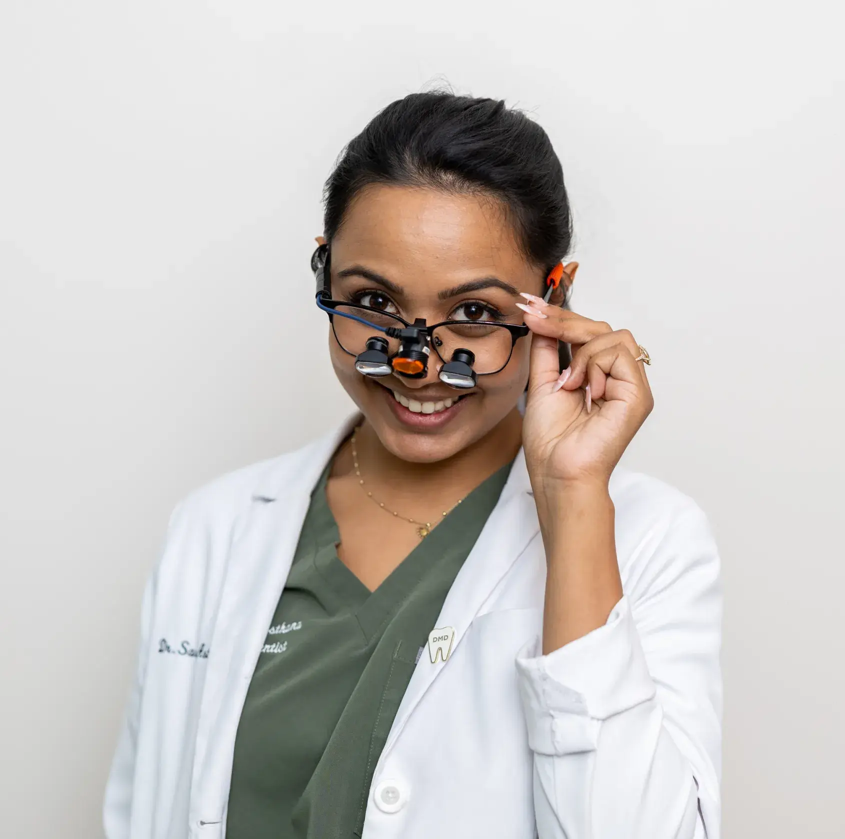 Smiling female dentist wearing magnification glasses and white coat with dental pins.