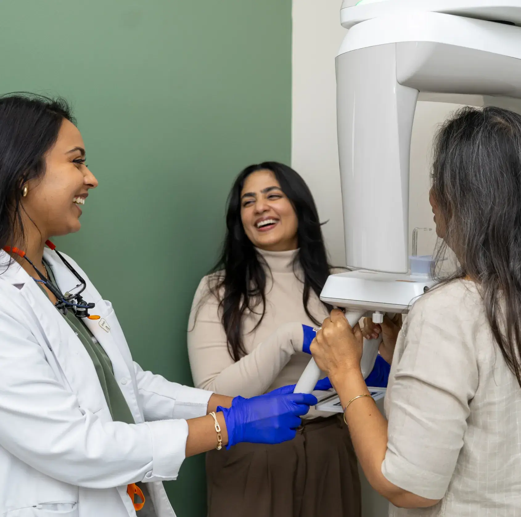 Dentist wearing blue gloves assisting a woman with a dental X-ray machine as another woman smiles in the background.