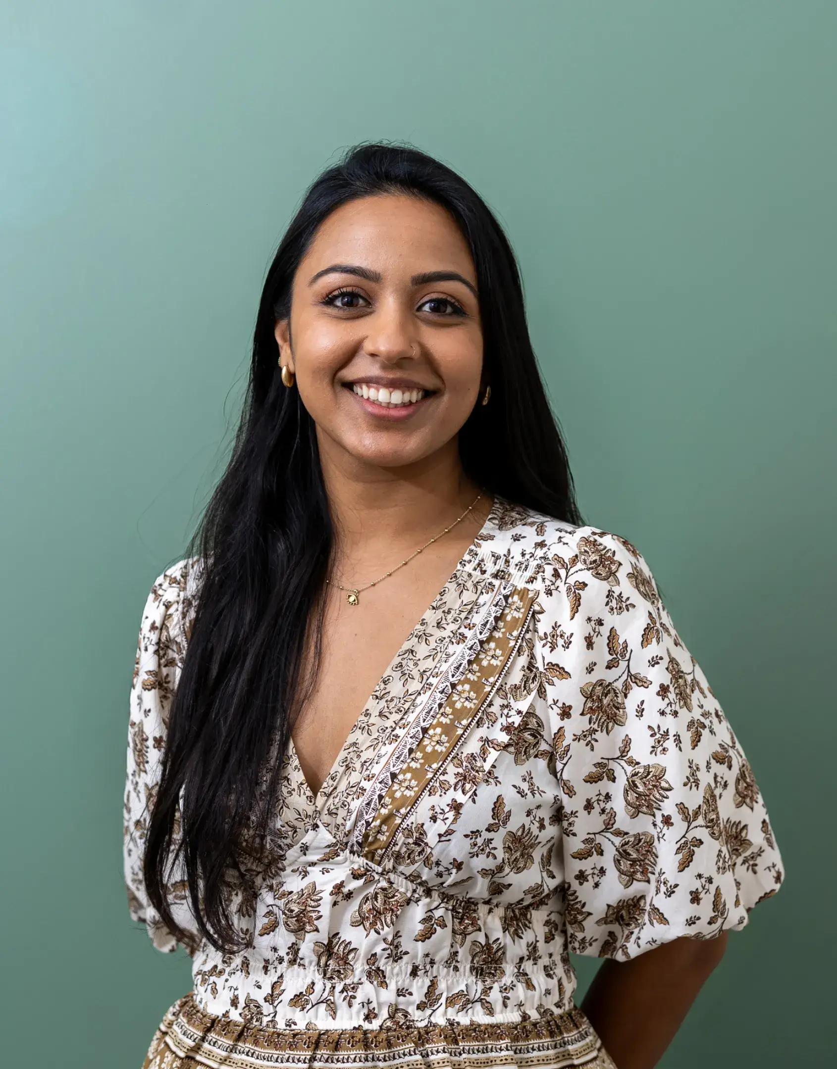 Smiling woman with long dark hair wearing a white and brown patterned dress against a green background.