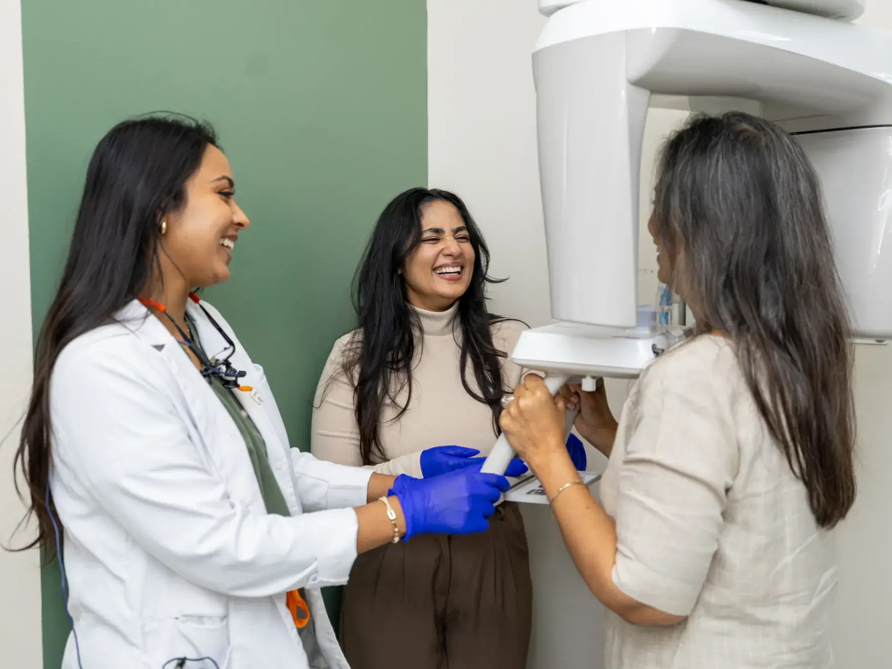 Two women assisting a smiling woman standing at a dental X-ray machine in a clinic.