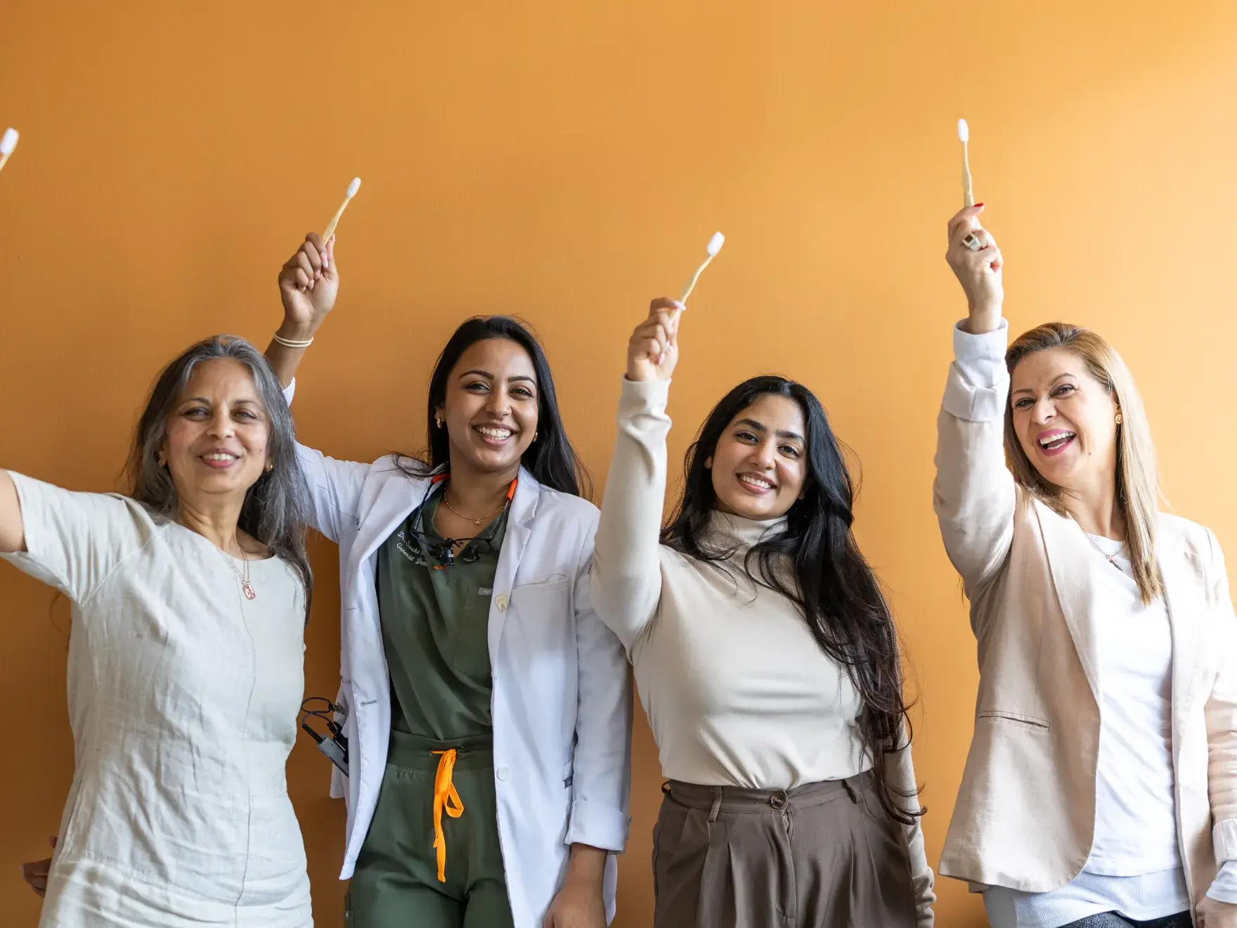 Four women smiling and holding up toothbrushes against an orange background.