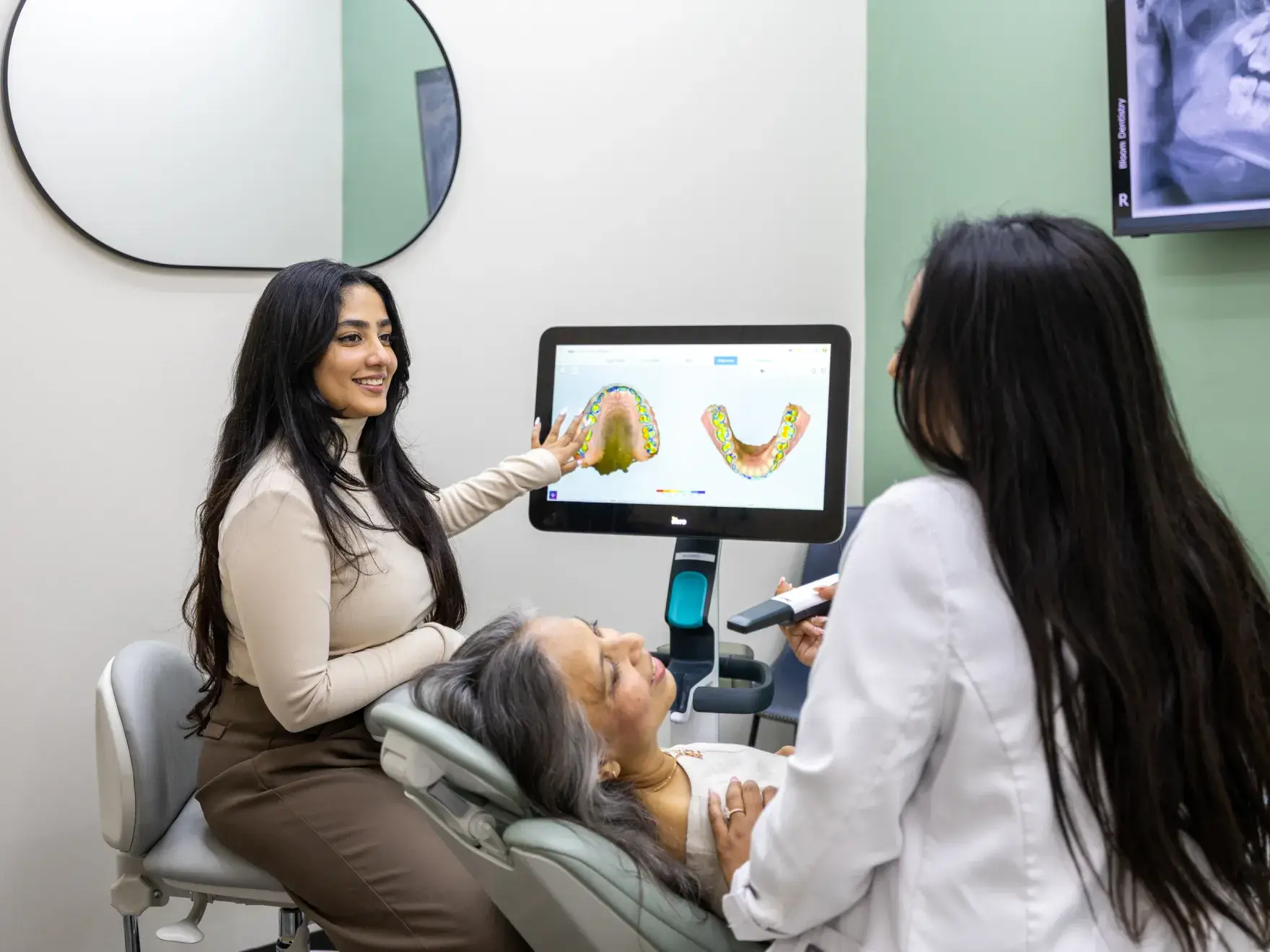 Dentist showing a dental scan on a monitor to a patient lying in the dental chair with another person explaining the image.