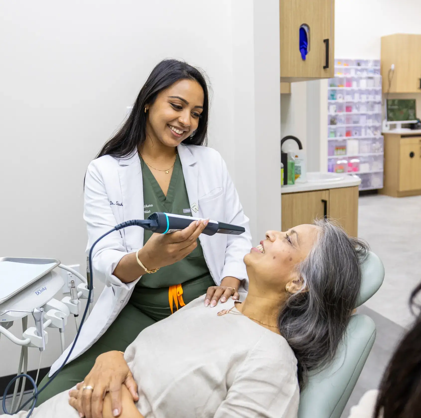 Dentist in white coat smiling and holding a dental scanning tool near a reclining female patient's mouth in a dental office.