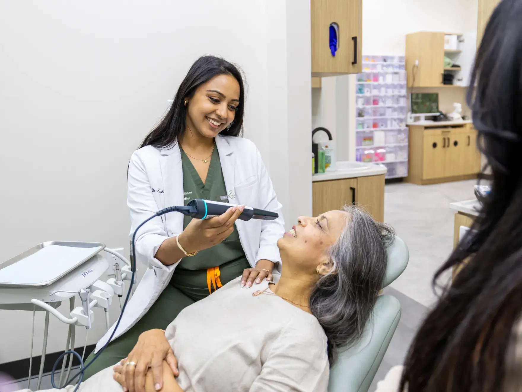 Dentist smiling and holding a dental scanner near a seated older woman in a dental clinic.