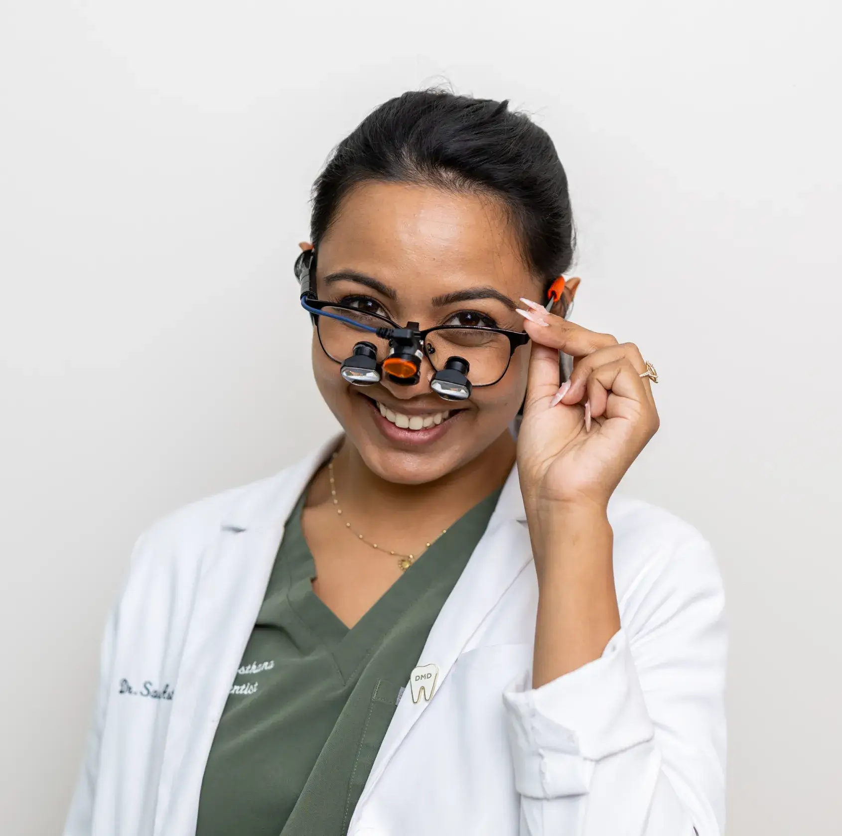 Smiling female dentist in white coat and dental loupes adjusting her glasses.