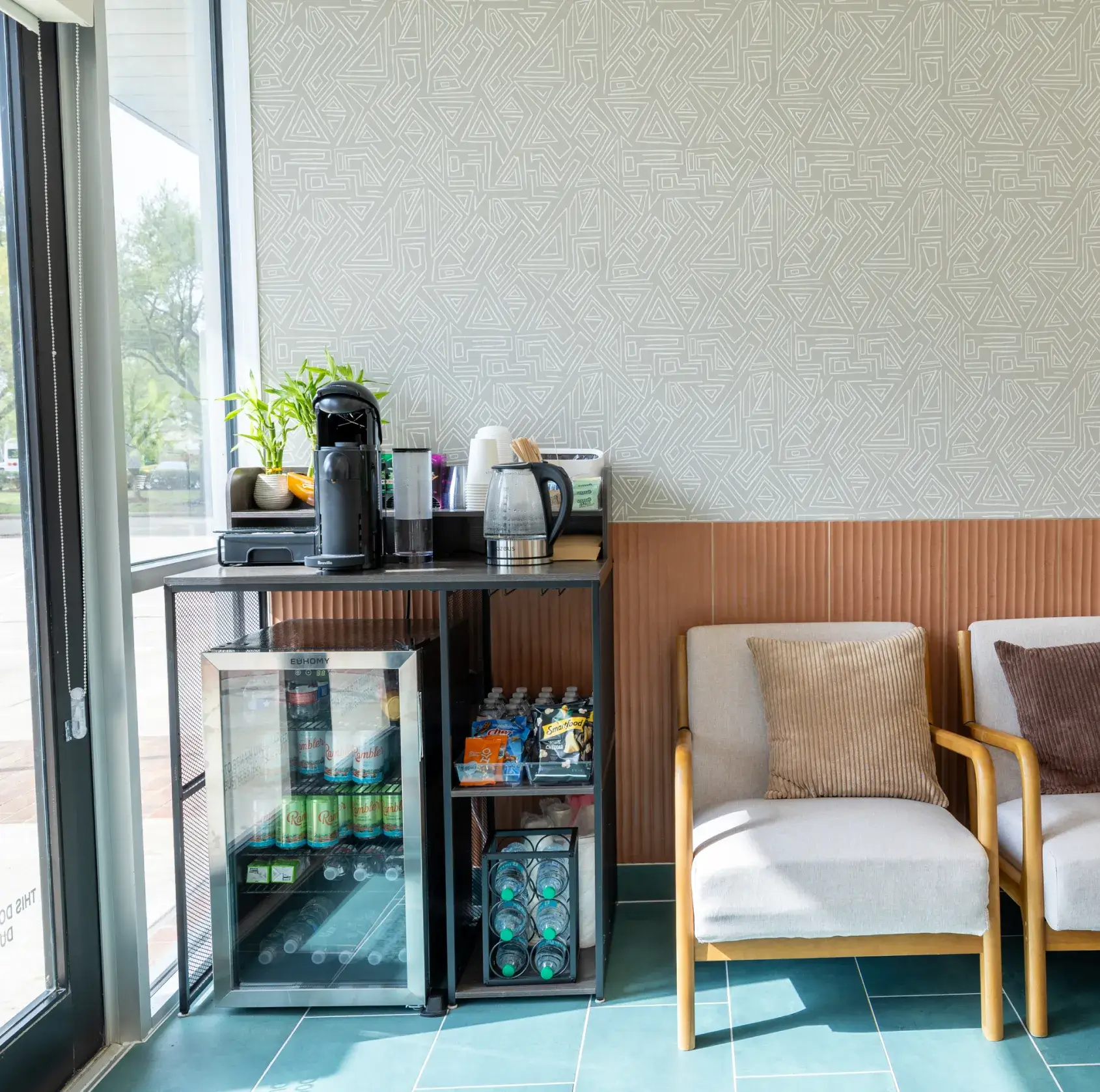 Coffee and beverage station with a small fridge, snack packs, coffee maker, and kettle next to two cushioned chairs with pillows in a bright room with patterned and wood-paneled walls.