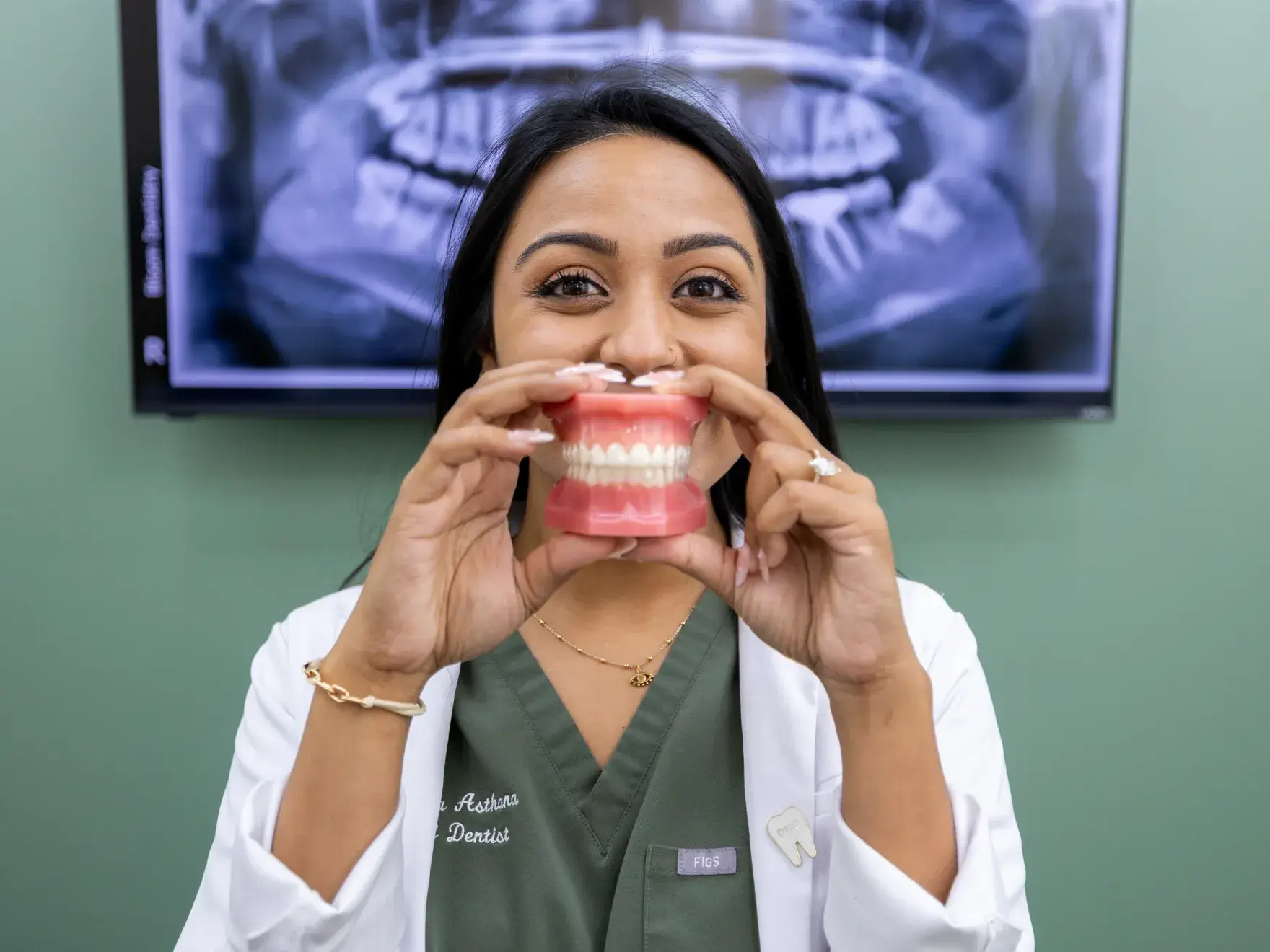 Dentist in green scrubs and white coat holding a dental model in front of her face with a dental X-ray on the screen behind her.