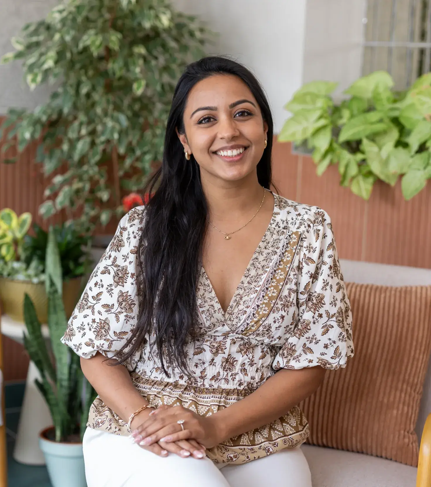 Smiling woman with long dark hair wearing a patterned blouse, sitting on a sofa with plants in the background.