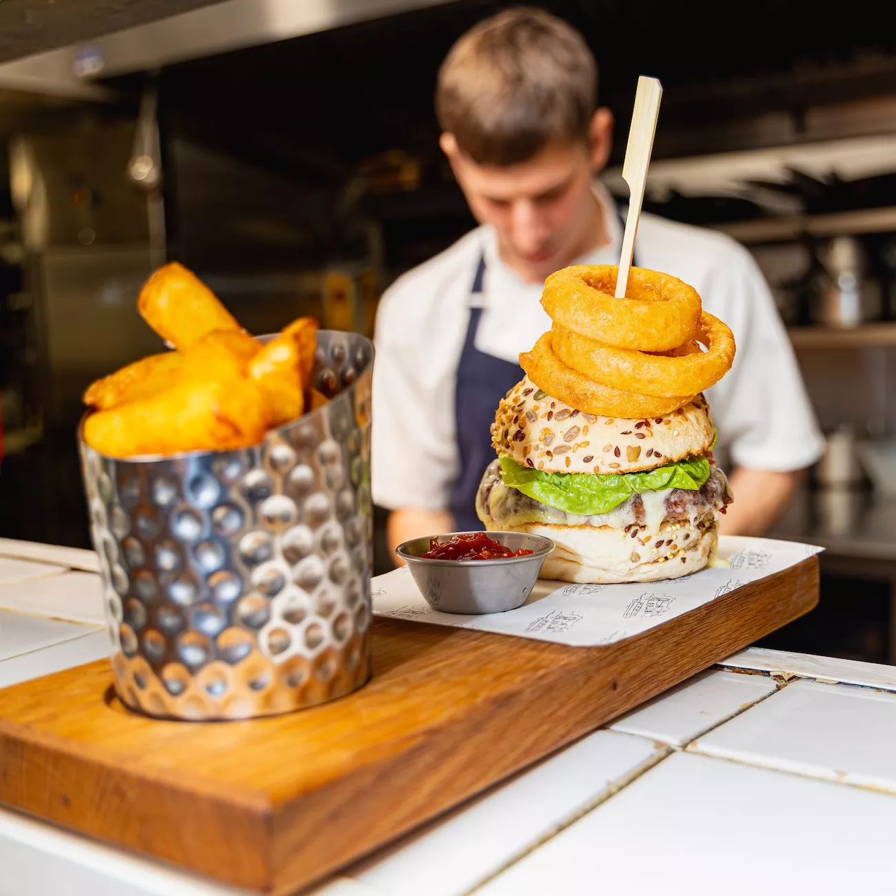 Chef at the pass behind a stacked cheeseburger and hand-cut chips in the kitchen of The Bridge Inn & Ferry in Offenham