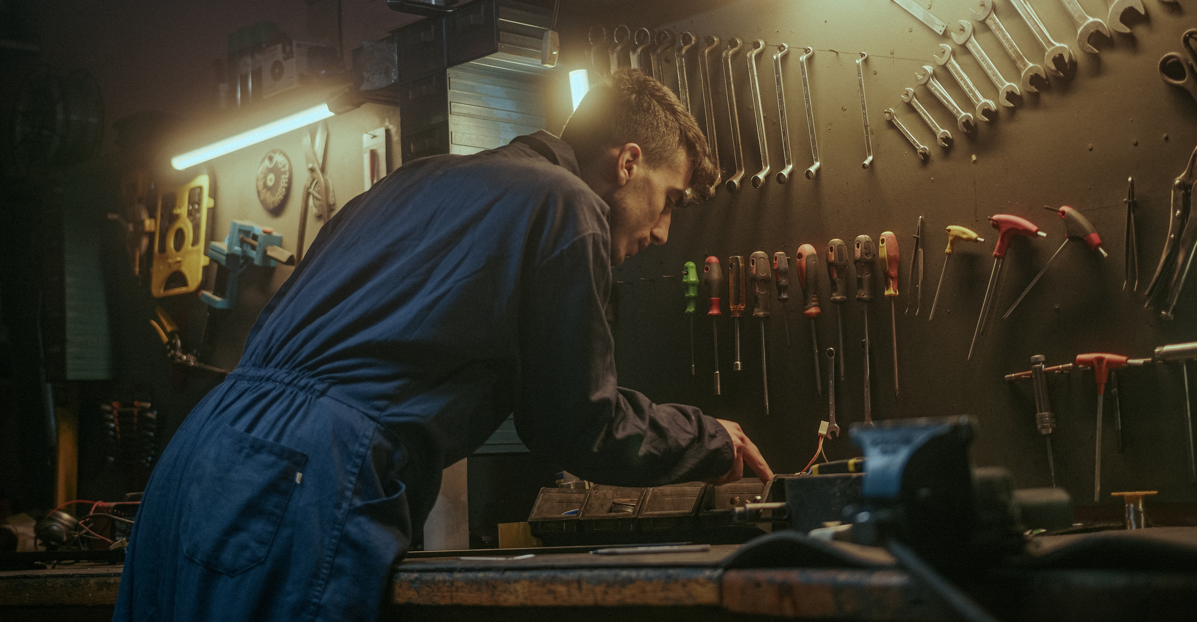 Man in blue coveralls leaning over a workbench in a dimly lit workshop with tools hanging on the wall.