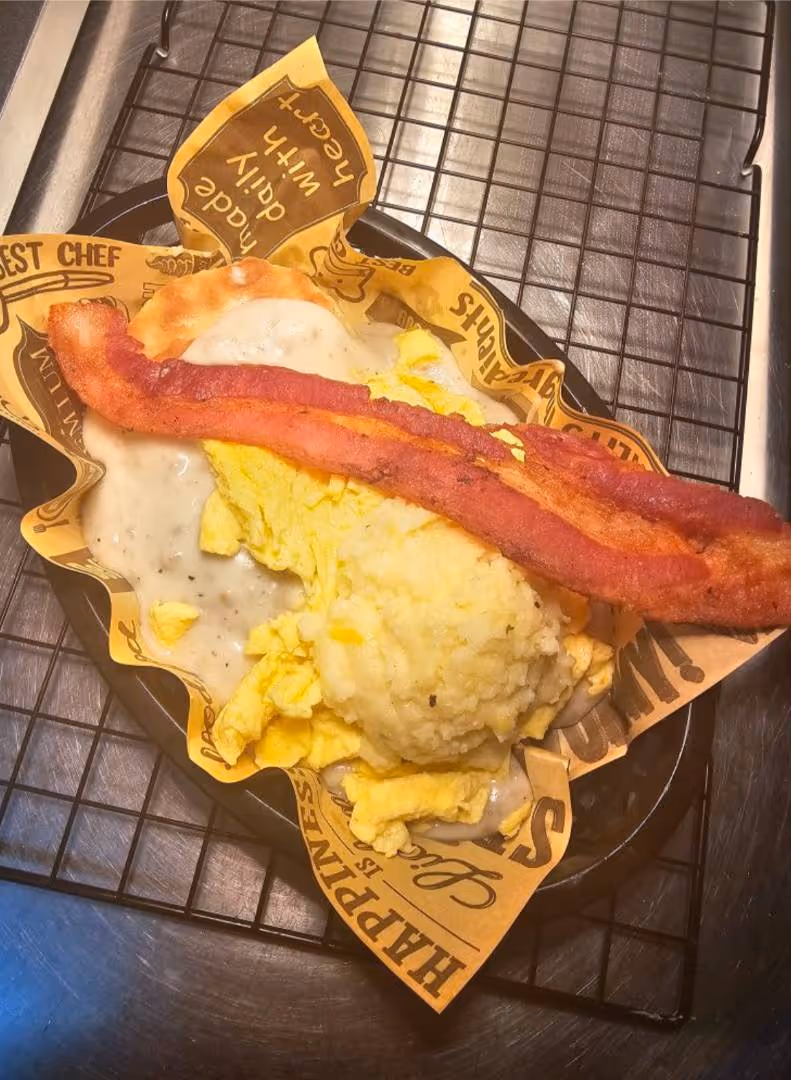 A plate with scrambled eggs, creamy gravy, mashed potatoes, and crispy bacon atop branded paper on a wire rack.