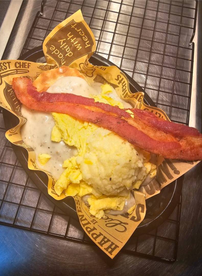 A plate with scrambled eggs, creamy gravy, mashed potatoes, and crispy bacon atop branded paper on a wire rack.