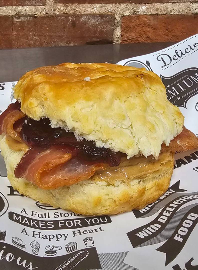A close-up of a sandwich on a printed wrapper. The sandwich features a flaky biscuit, crispy bacon, peanut butter, and dark jam.